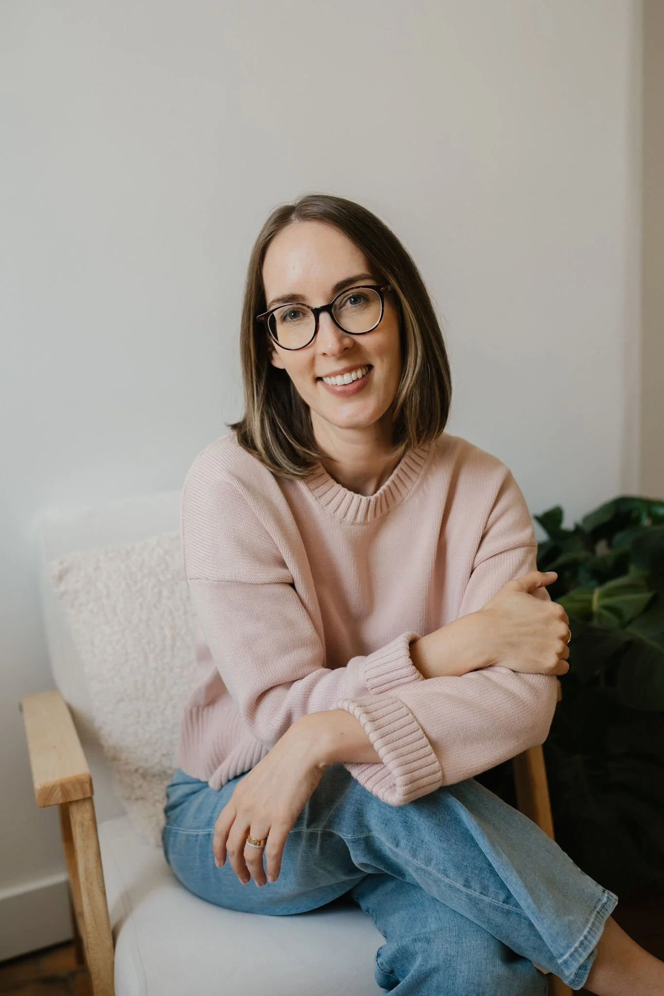 A woman with shoulder-length brown hair, glasses, and a pink sweater is sitting on a wooden chair with her arms crossed, smiling. There is a green plant in the background and a white wall.