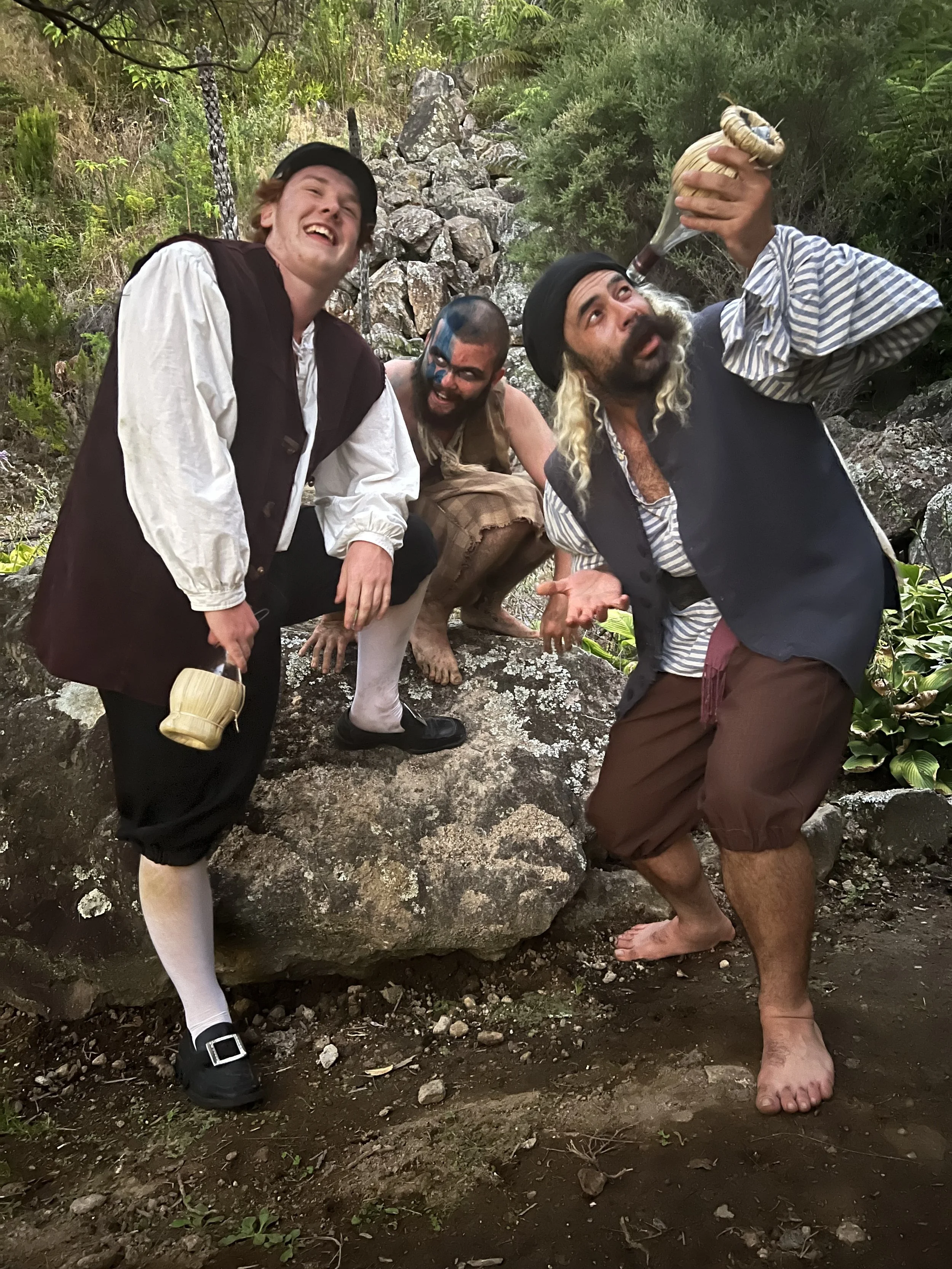 Trinculo, Stephano and Caliban from The Tempest in 2024 production. Three men dressed as pirates in a forest, one drinking from a bottle, one crouching behind him, and the third holding a mug, all smiling.