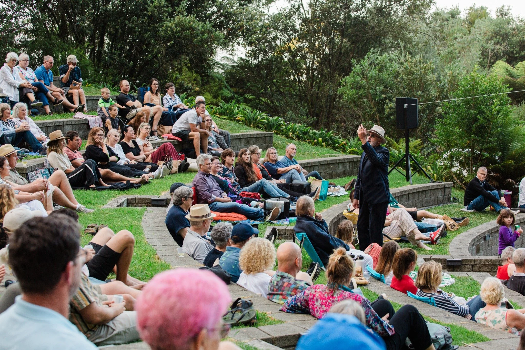 Prince from Romeo and Juliet performing in front of a seated audience in the Te Puna Quarry Park Amphitheatre on terraced grass seats surrounded by trees.
