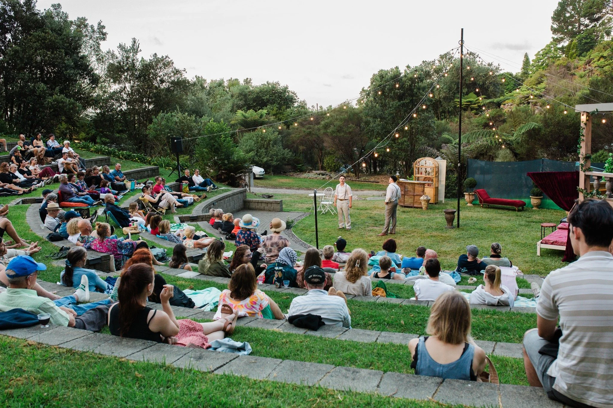 An outdoor theater performance at Te Puna Quarry Park Amphitheatre with an audience seated on tiered steps and on the grass, watching two actors on stage under string lights, surrounded by trees and greenery.
