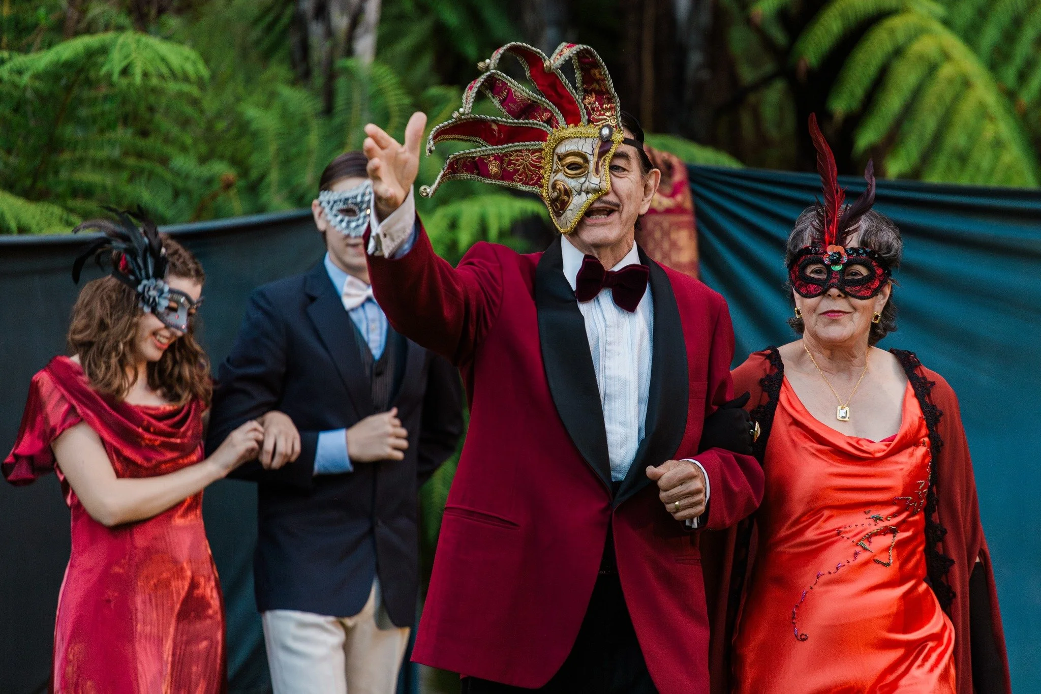 Romeo and Juliet cast of the Capulet family. Capulet, lady Capulet and Juliet at a masquerade party wearing elaborate masks and fancy clothing, Capulet in red jacket and mask in the center.