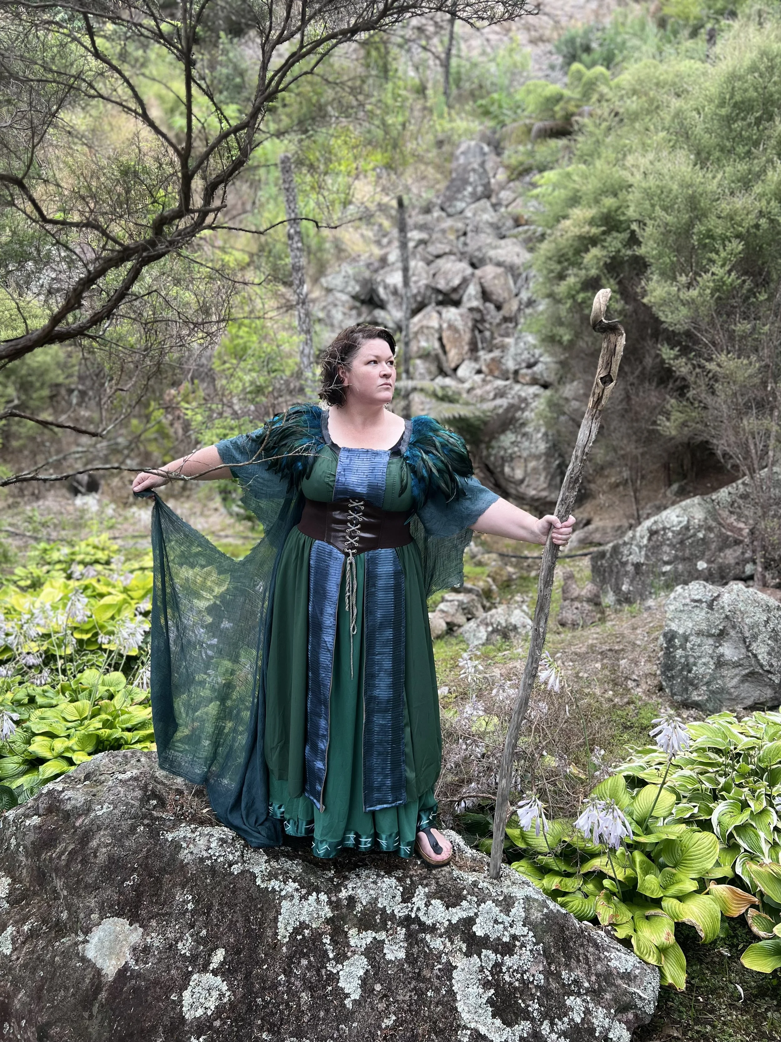 Gayle Spence as Prospero in 'The Tempest' in 2024 at Te Puna Quarry Park Amphitheatre dressed in fantasy costume standing on a large rock in a forested area with rocks and green foliage, holding a wooden staff.