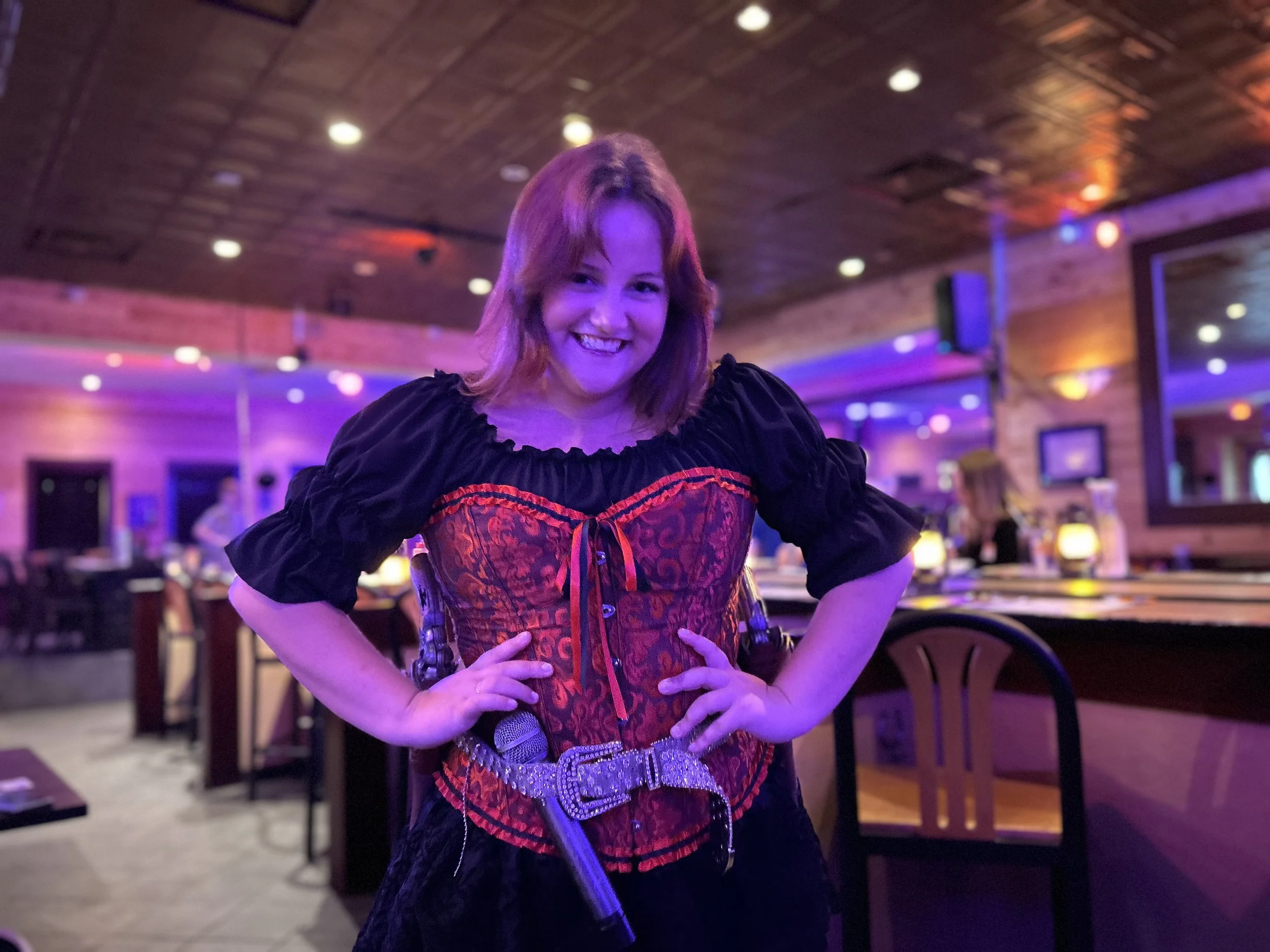 A young woman with shoulder-length red hair smiles at the camera in a dimly lit bar or restaurant. She wears a black and red lace corset with black puffed sleeves, and has her hands on her hips. The background features wooden walls, ceiling, and tables with warm lighting and purple accents.