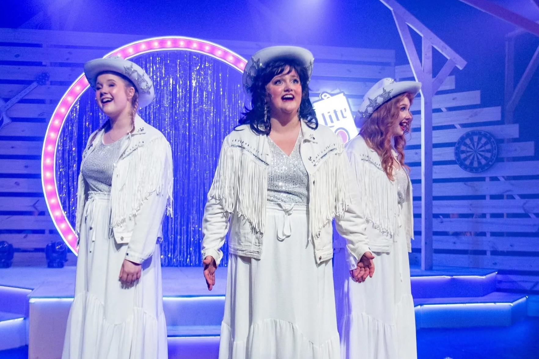 Three women dressed in white Western-style outfits and hats, standing hand-in-hand on a stage decorated with a retro or country theme, bright lights, and a background with a heart-shaped outline and a dartboard.