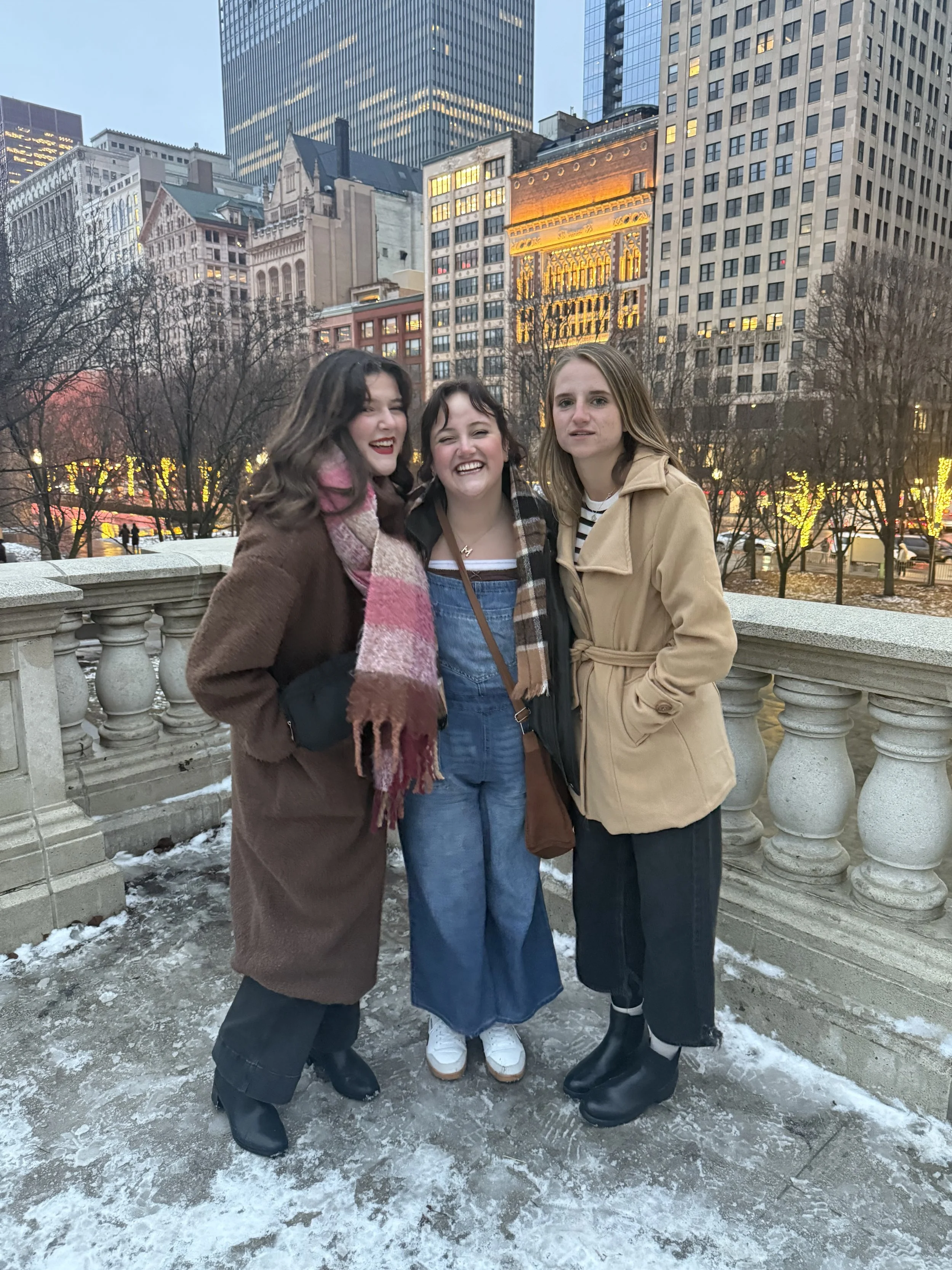 Three young women standing on a snowy outdoor balcony overlooking a cityscape with tall buildings and illuminated windows in the evening, smiling and posing for the photo.