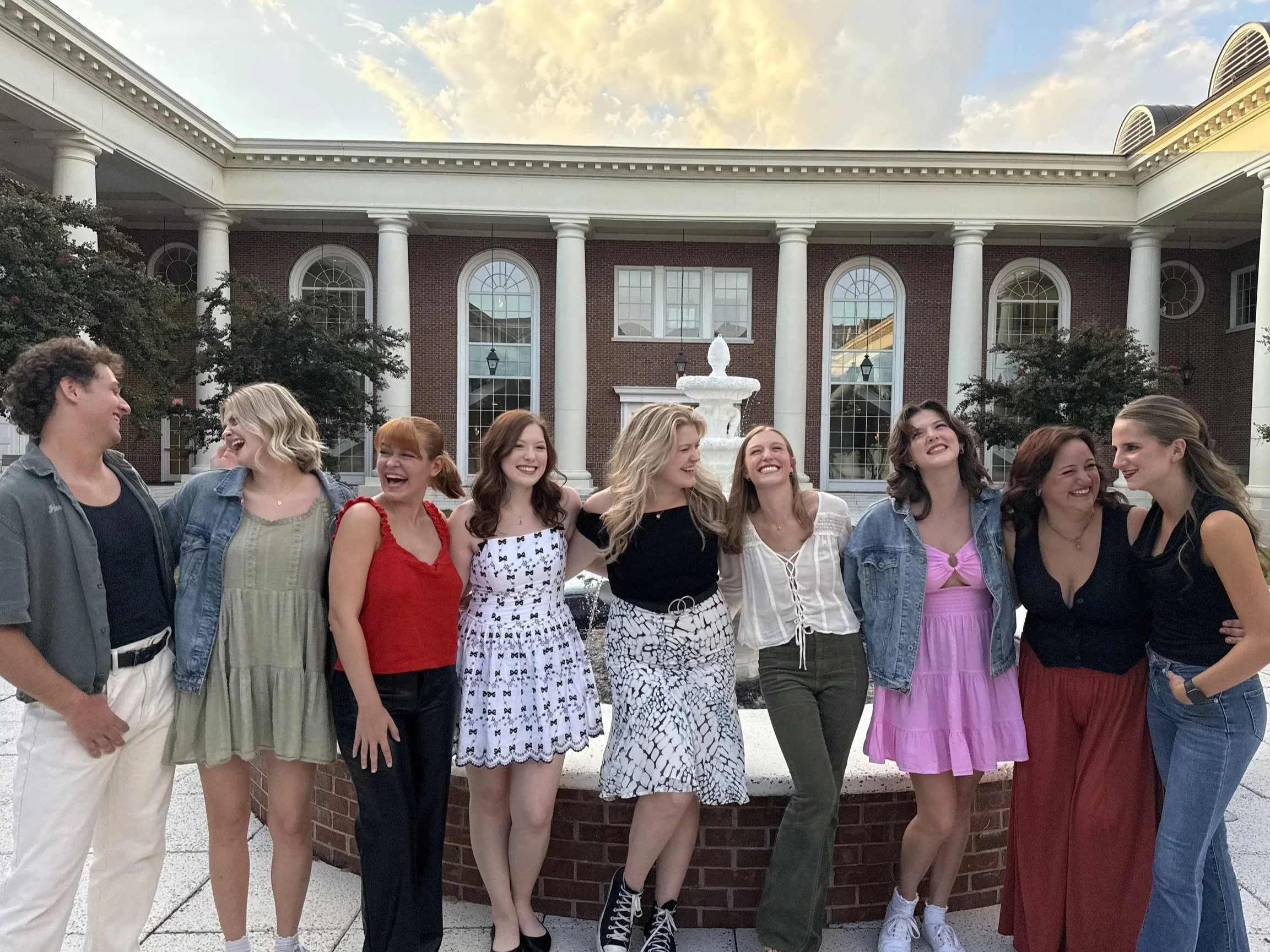 A group of nine young women laughing and smiling together outside in front of a fountain and a building with large columns.