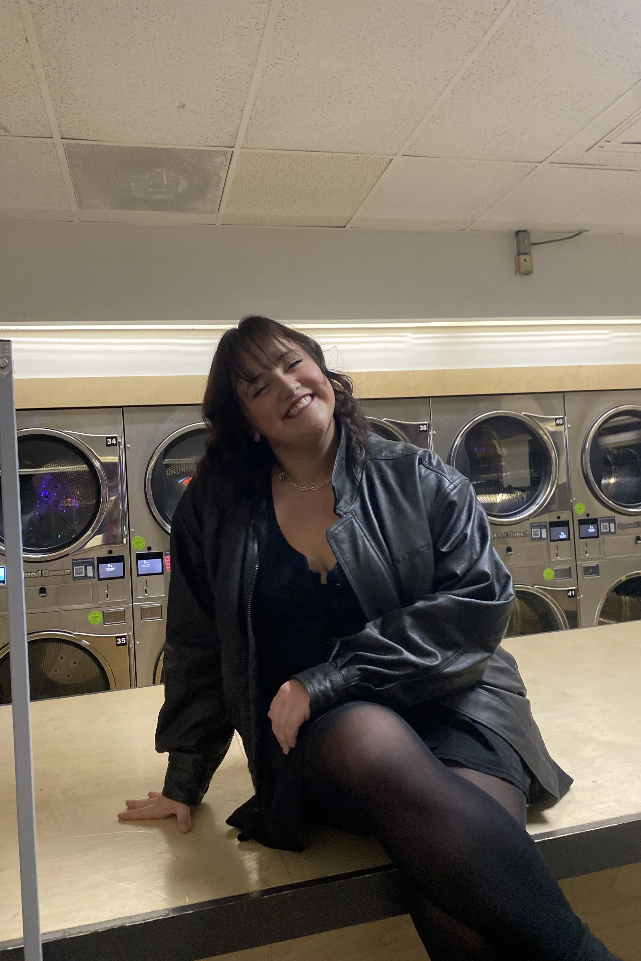 A woman sitting on a counter in a laundromat, smiling, wearing a black dress, black tights, and a black leather jacket.