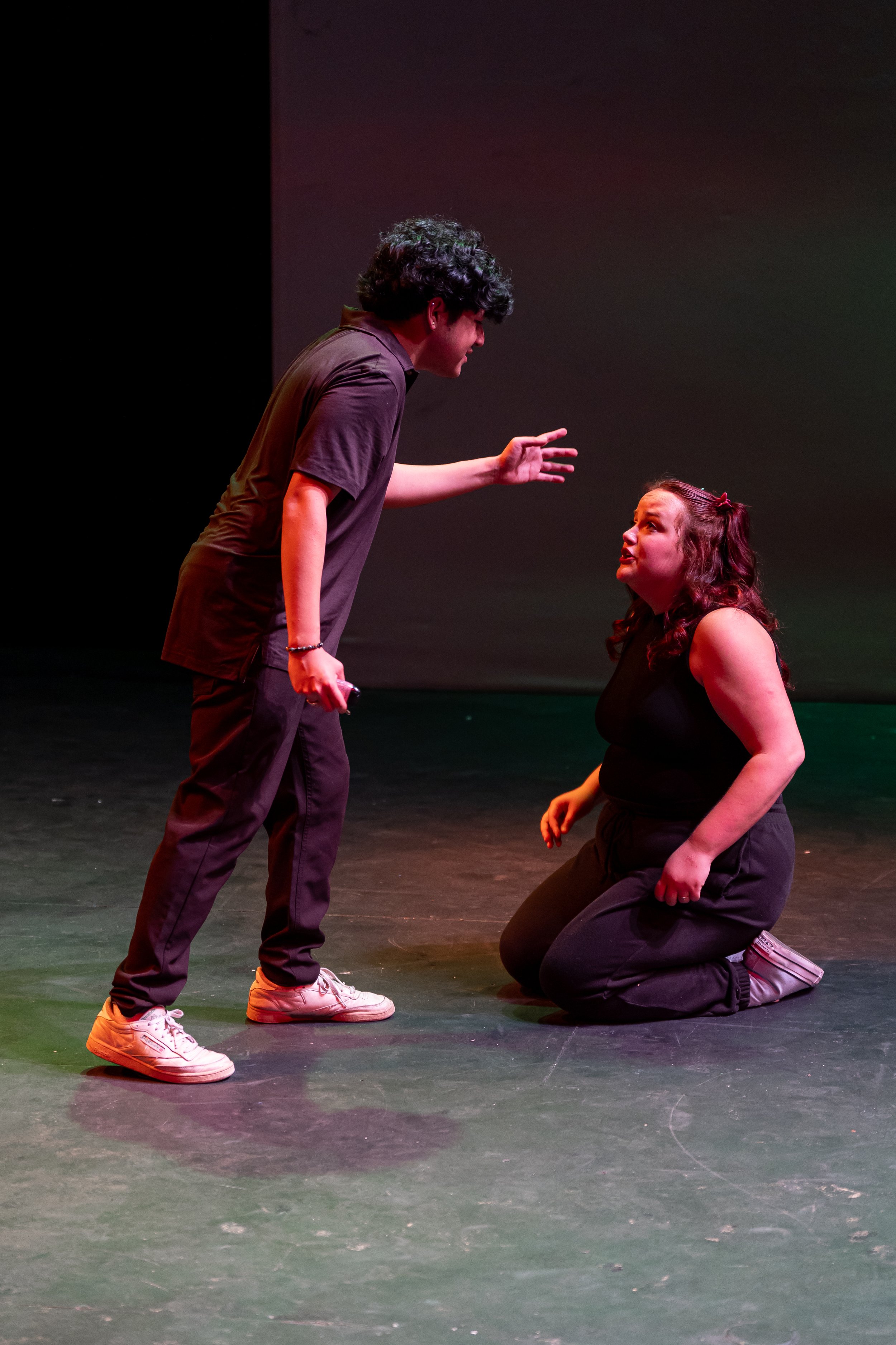 Two young actors performing on stage; one is standing with a hand raised, the other is kneeling, looking up at the standing actor, in a scene that appears emotional.