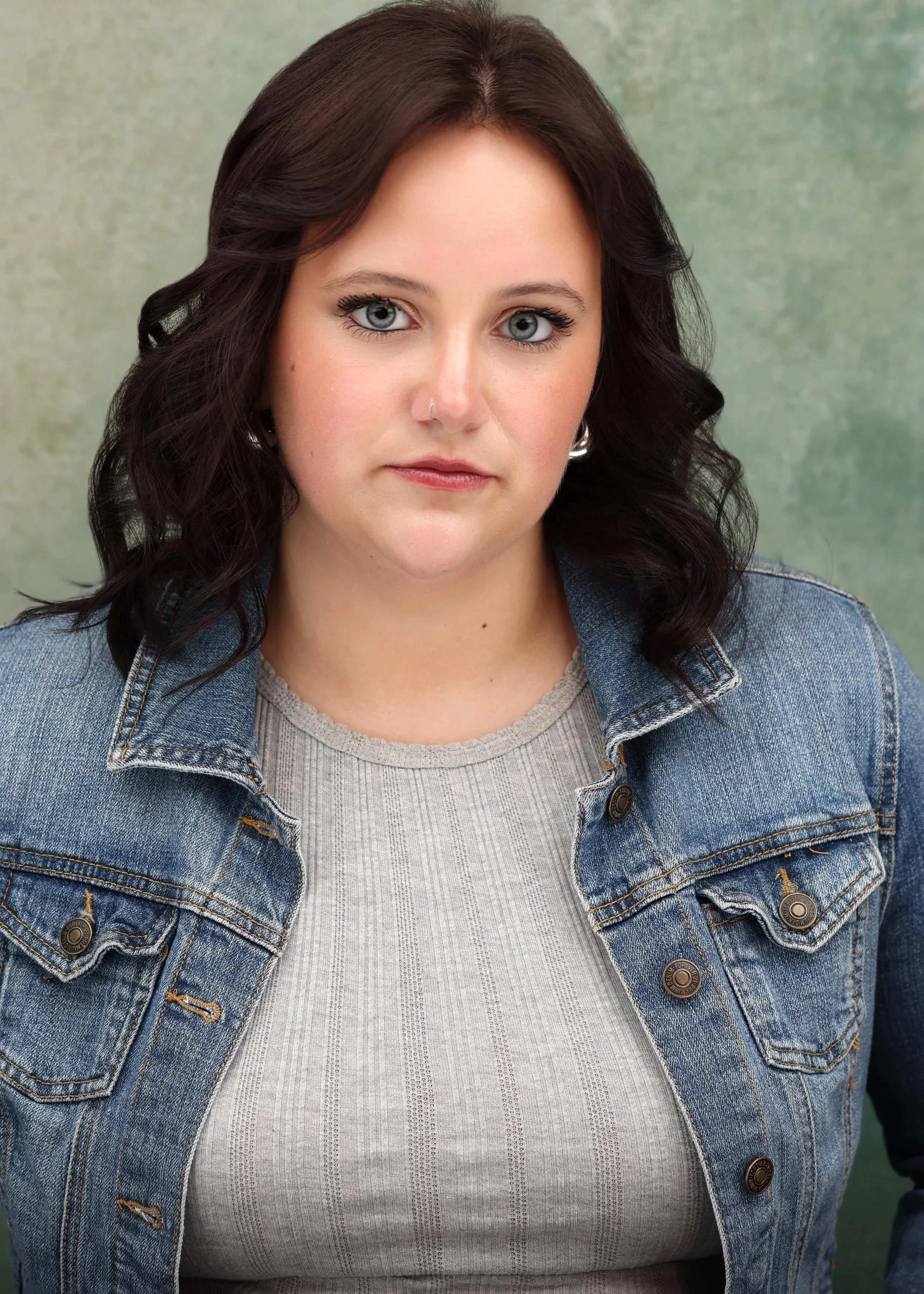 A young woman with dark wavy hair, blue eyes, wearing a light gray top and denim jacket, posed against a muted green background.