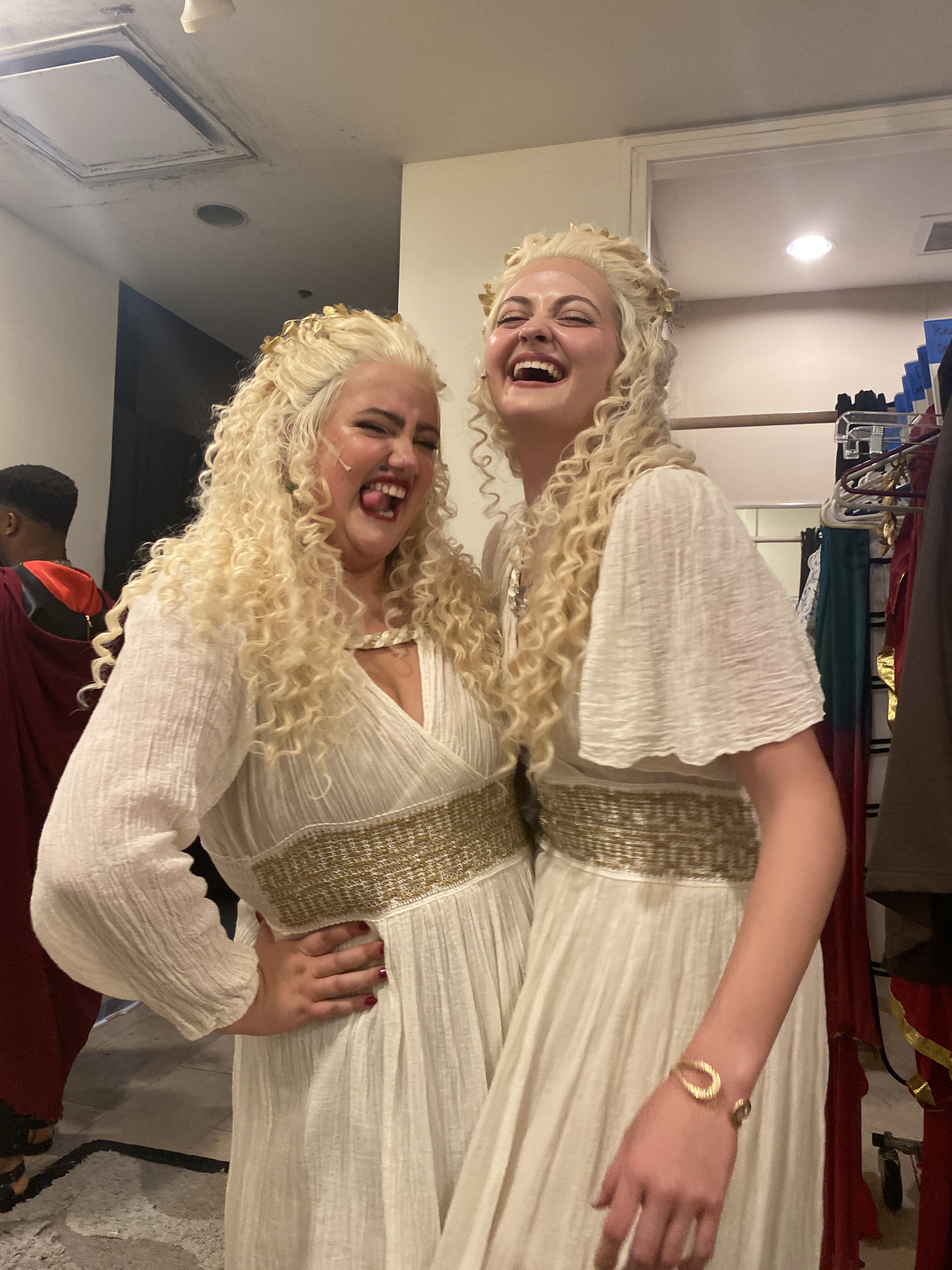 Two women dressed in cream-colored dresses with gold embellishments laughing and posing together indoors.