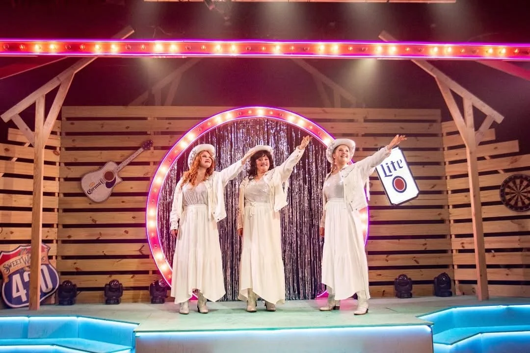 Three women dressed in white long dresses and white hats performing on stage with retro decor, including signs for Corona beer and Miller Lite, a guitar, and a dartboard, in a rustic wooden setting.