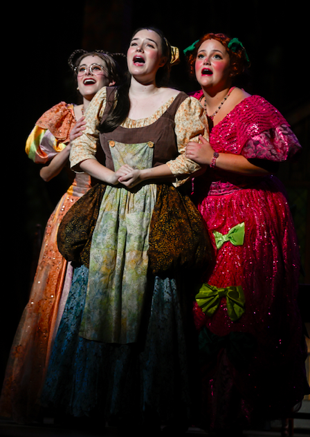 Three women dressed in colorful, vintage-style dresses perform on stage, appearing emotional, with the center woman crying and the two others comforting her.