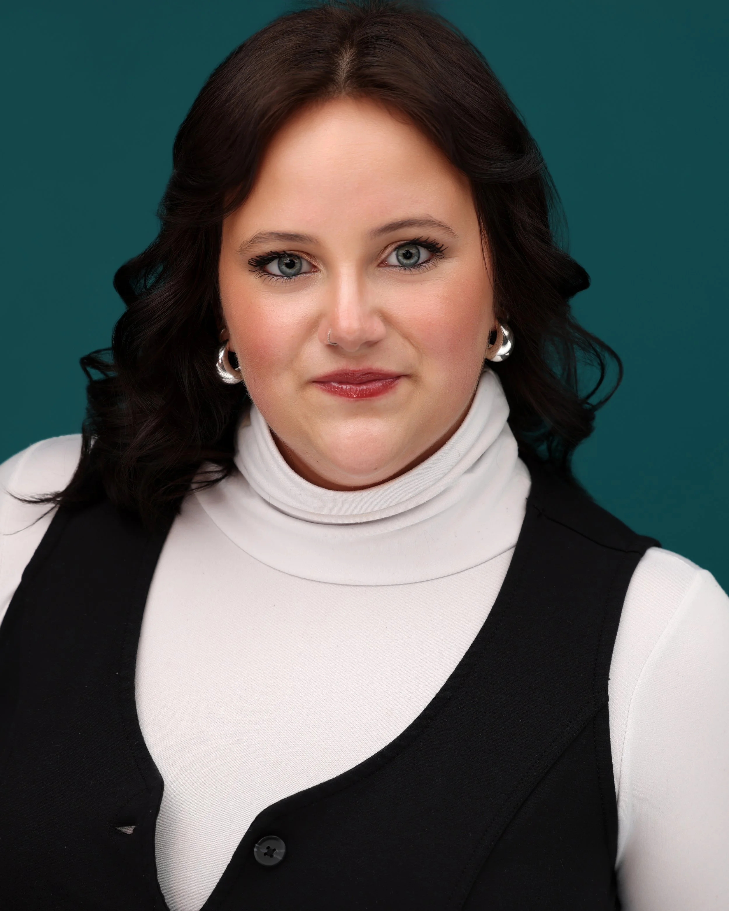 A woman with dark wavy hair, blue eyes, wearing a white turtleneck, black vest, earrings, and makeup, posing against a teal background.
