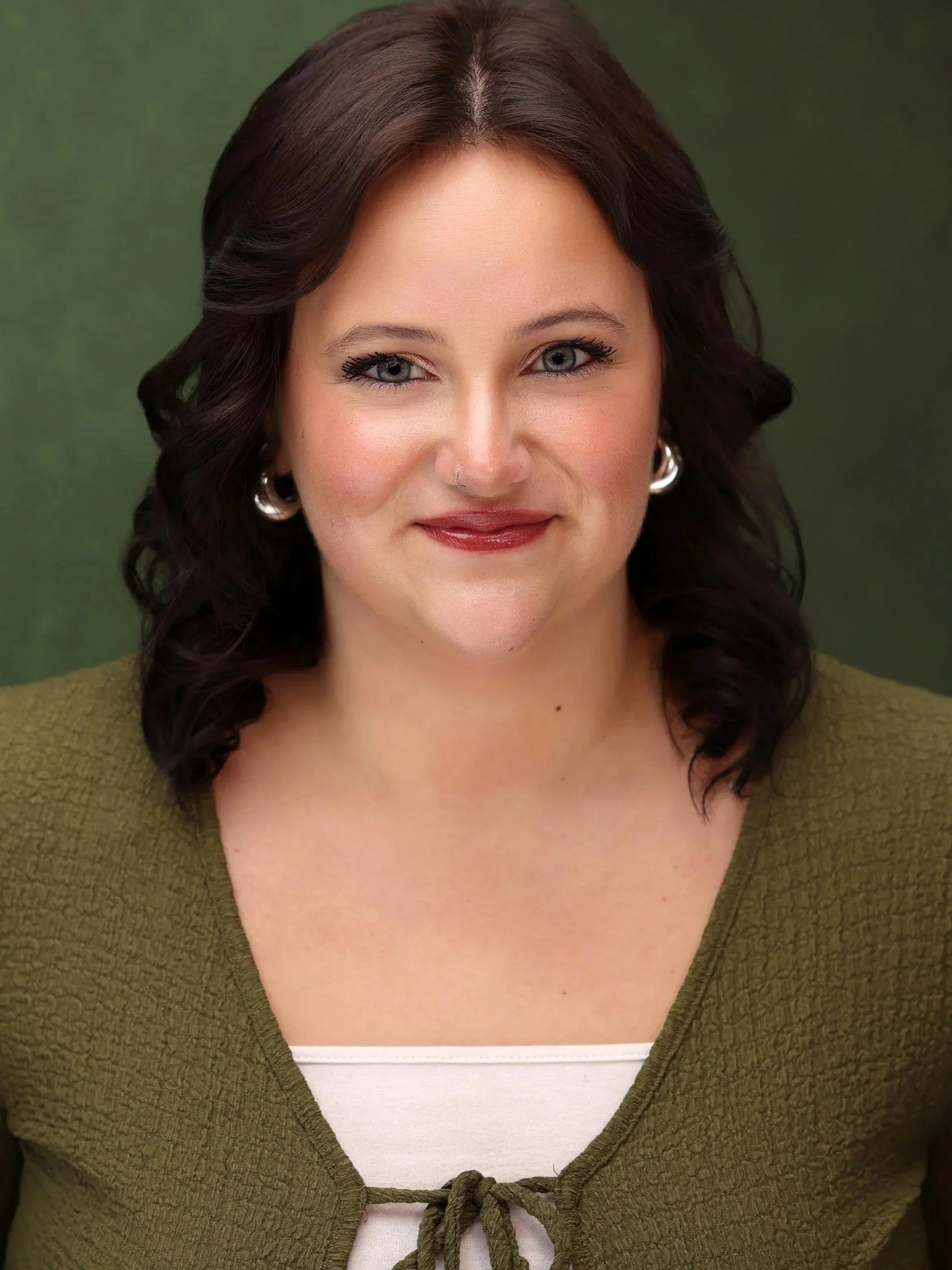 A woman with wavy dark brown hair, light skin, wearing pearl earrings and a nose ring, smiles in front of a green background, dressed in an olive green top with a white undershirt.