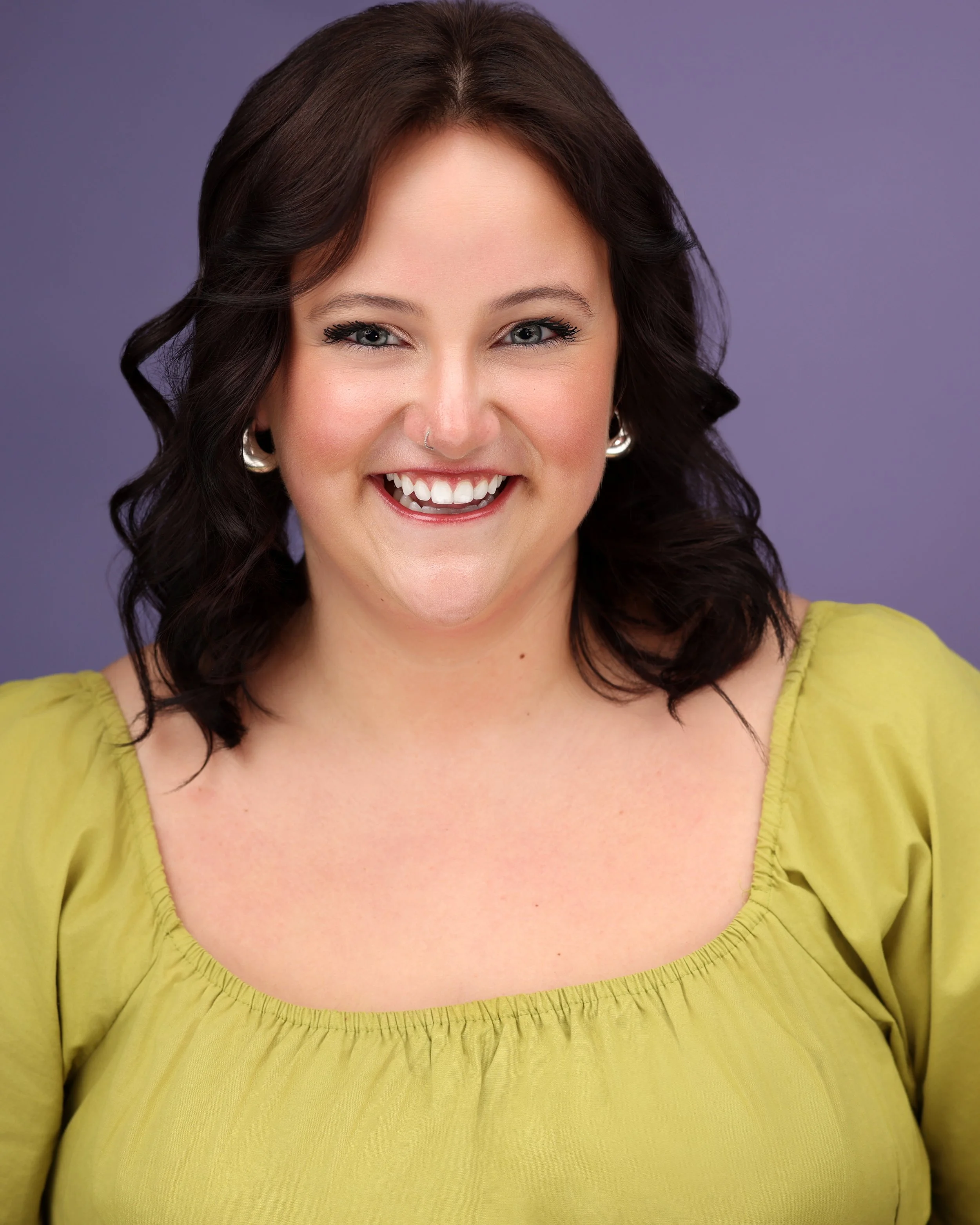 A woman with dark brown, wavy hair, light skin, wearing a yellow top, smiling with white teeth, against a purple background.