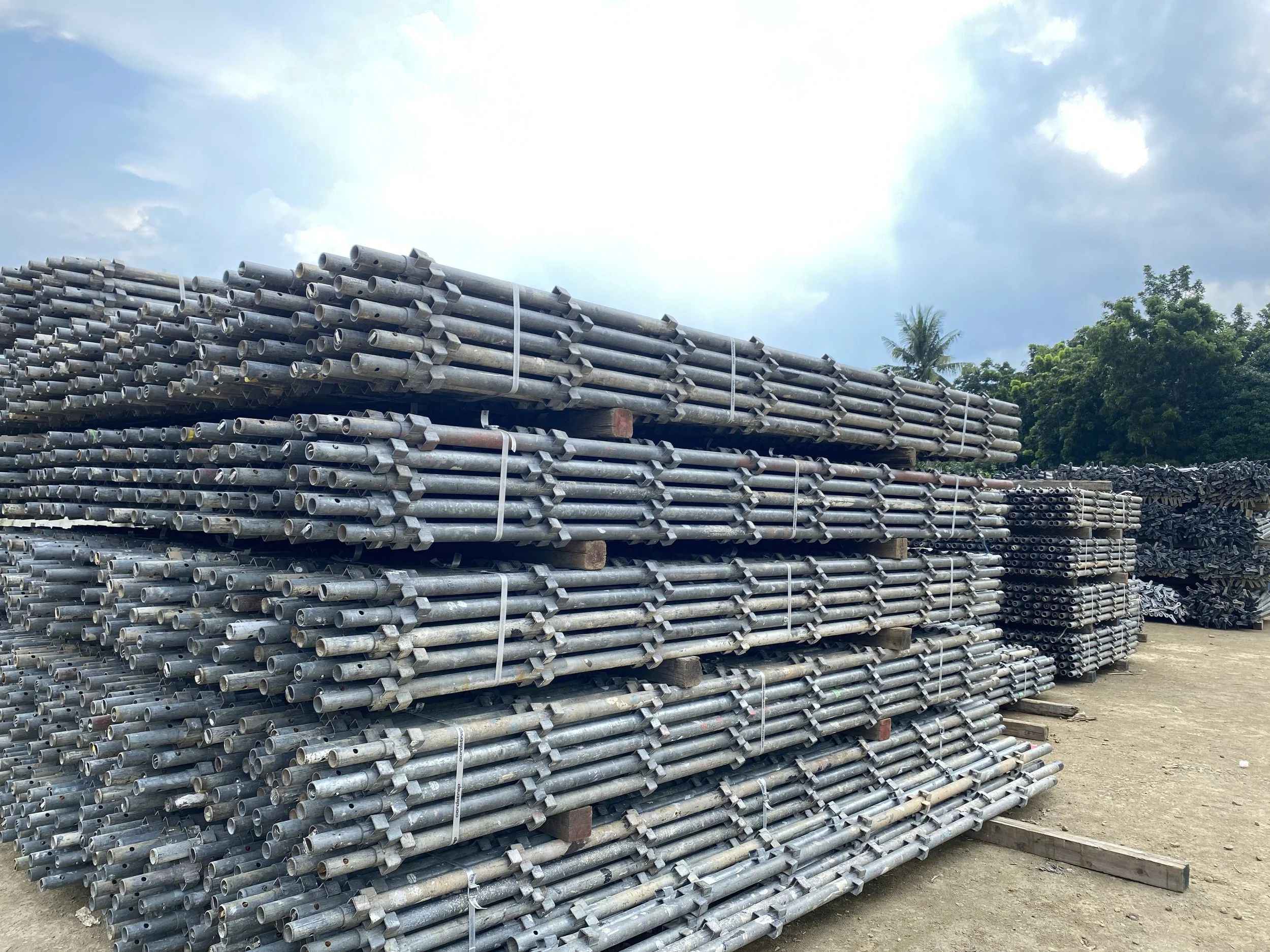 Stacks of metal pipes and scaffolding materials on a construction site with trees and cloudy sky in the background.