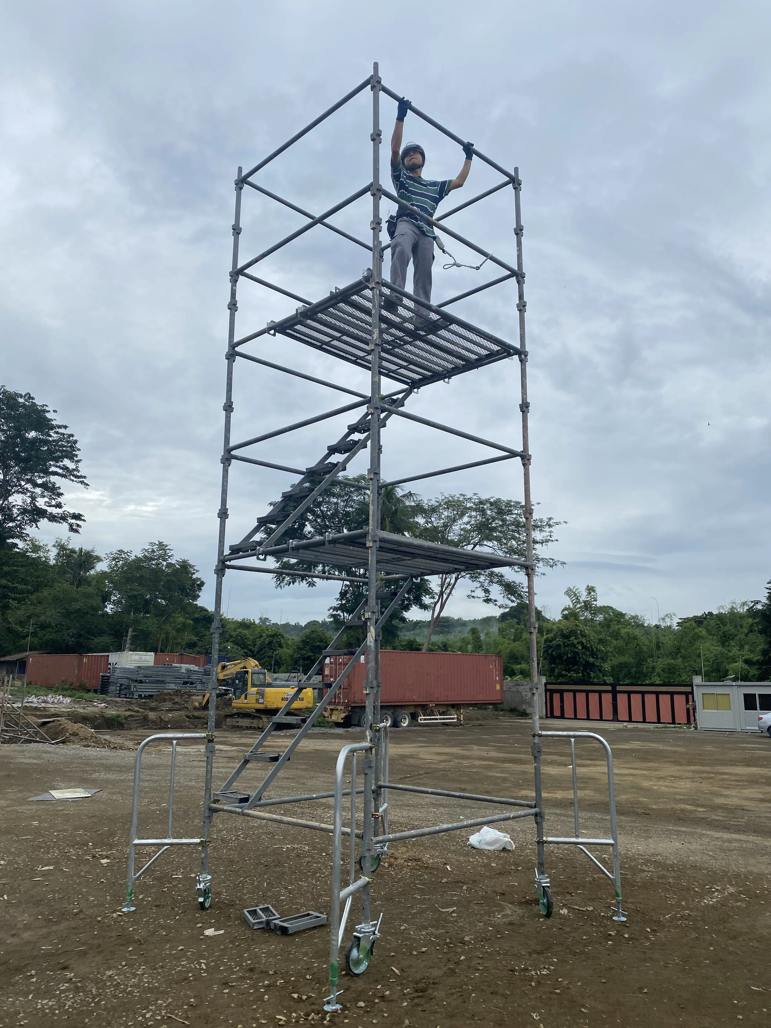 A man wearing a striped shirt, gray pants, and safety gloves standing on a metal scaffold at a construction site, holding onto the top of the scaffold structure.