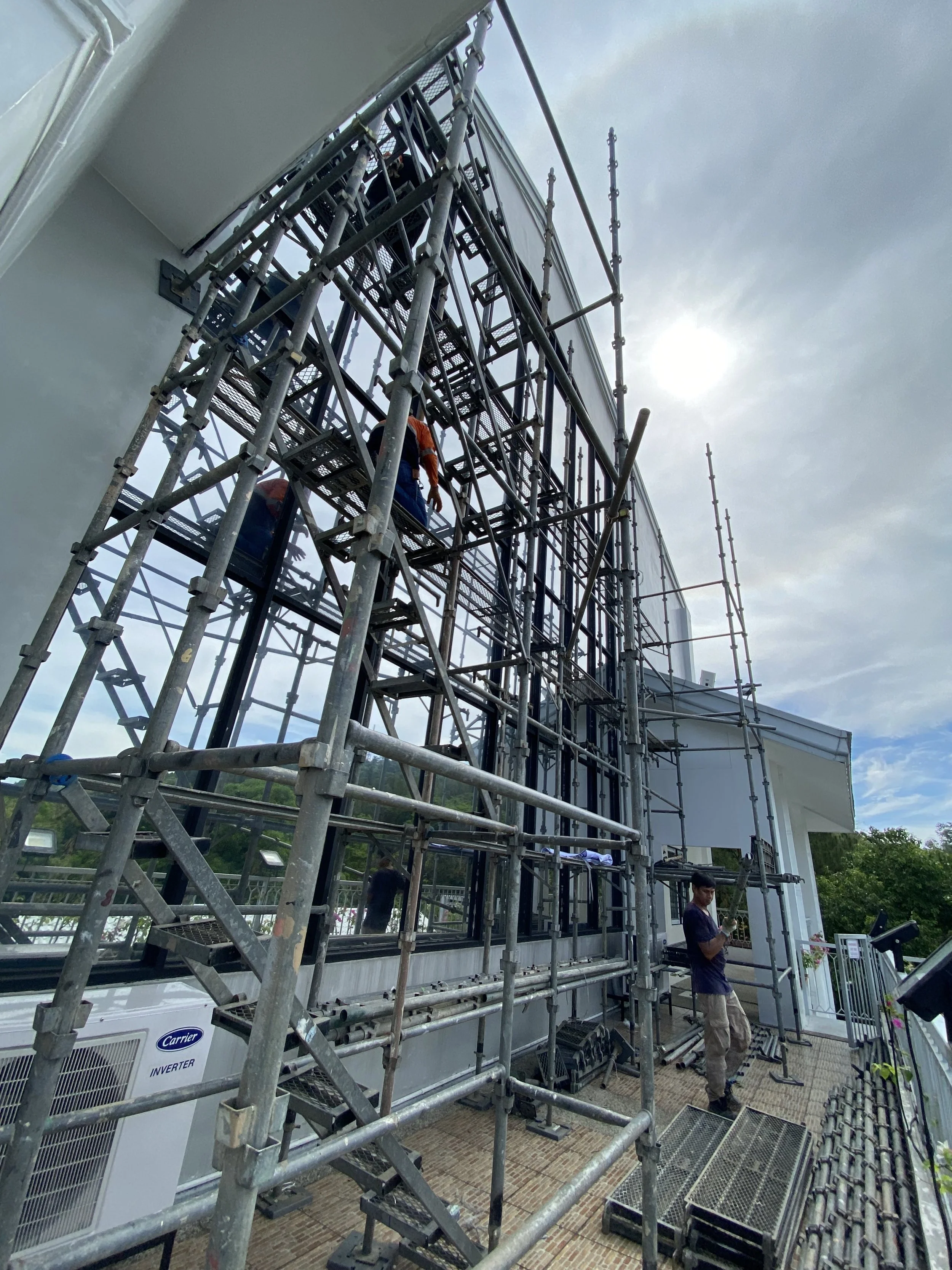 Construction workers on scaffolding outside a building with glass walls, with the sky and sun visible in the background.