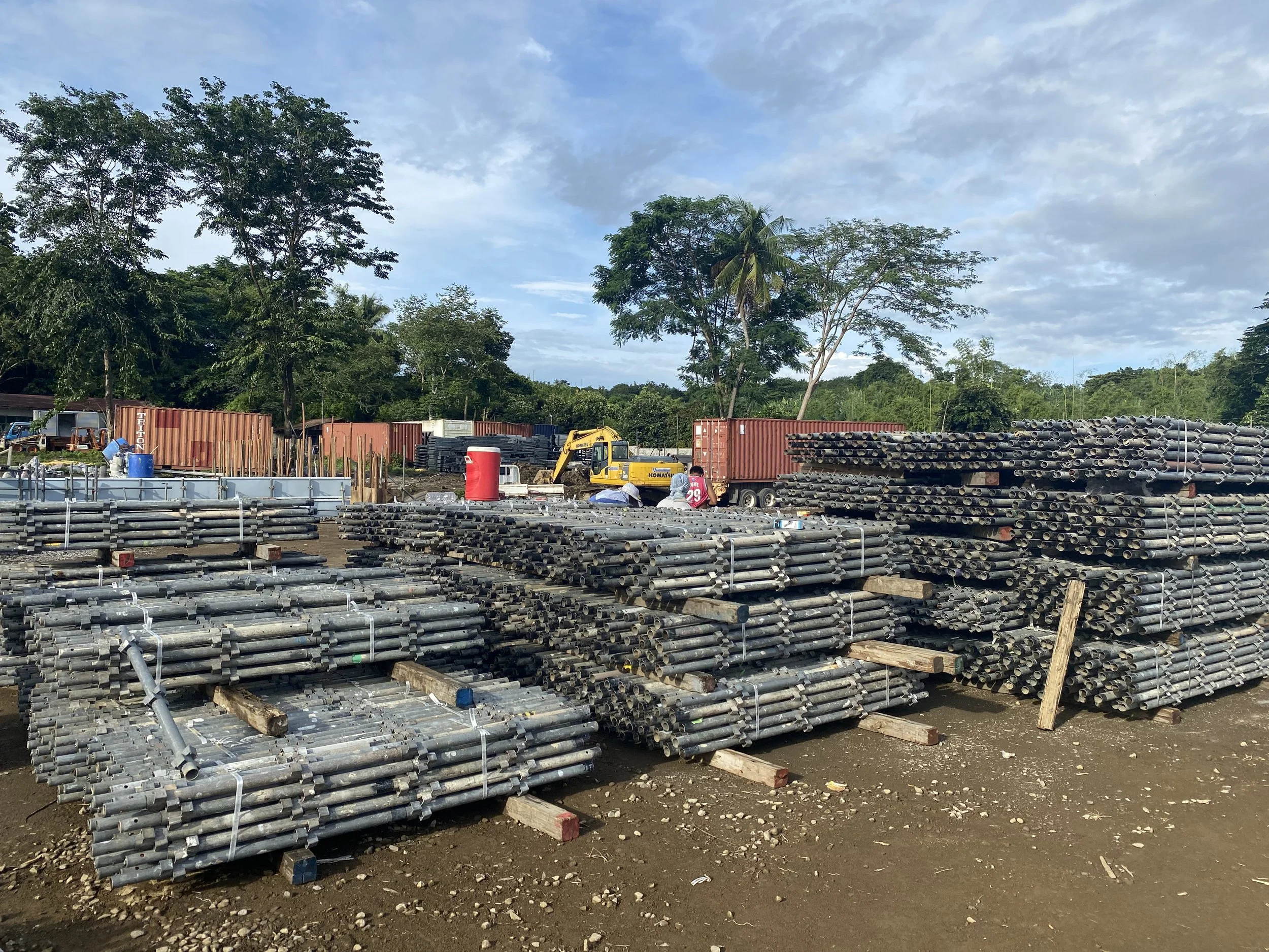 Stacks of metal pipes piled on wooden supports at a construction site with trees and shipping containers in the background under a partly cloudy sky.