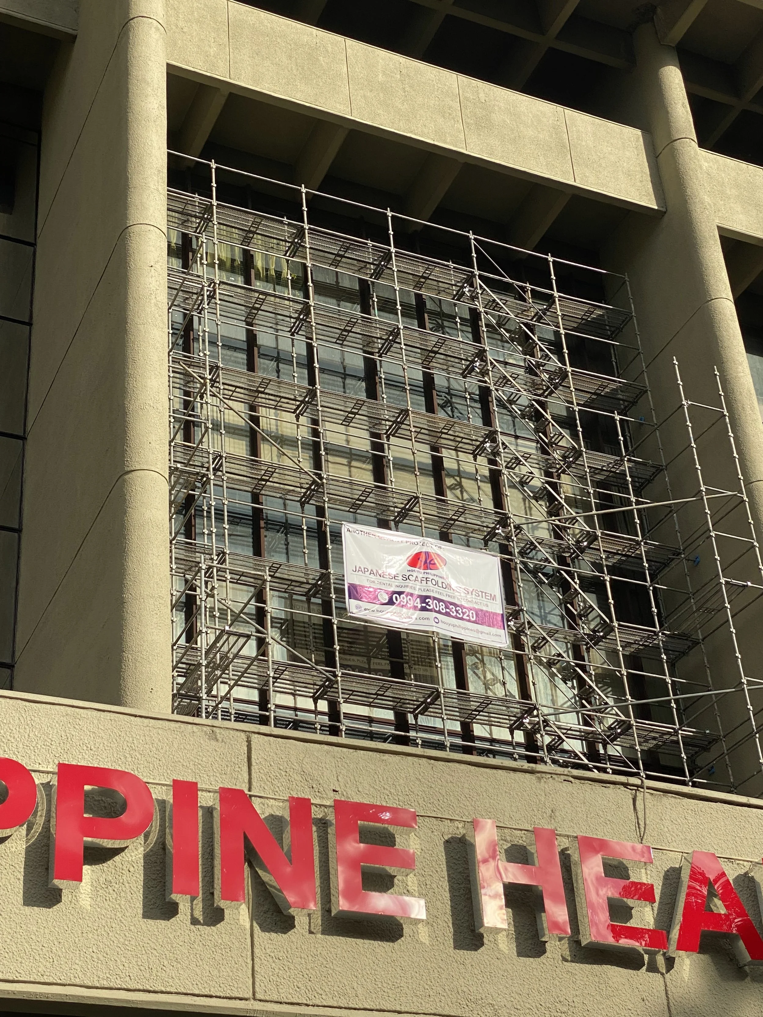 Construction scaffolding in front of a multi-story building with large glass windows and a banner advertising a Japanese scaffolding system, partially visible red sign reading 'PINE HEA'.