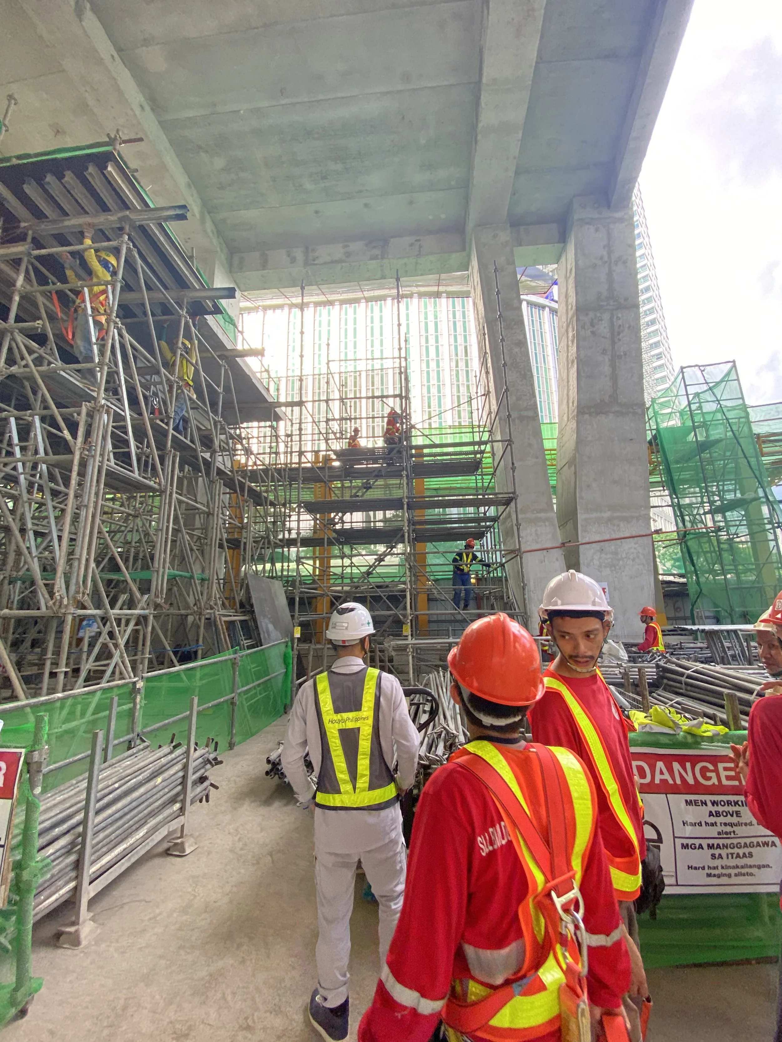 Construction site with workers wearing safety helmets and vests, scaffolding, concrete supports, and a partially completed building under construction.