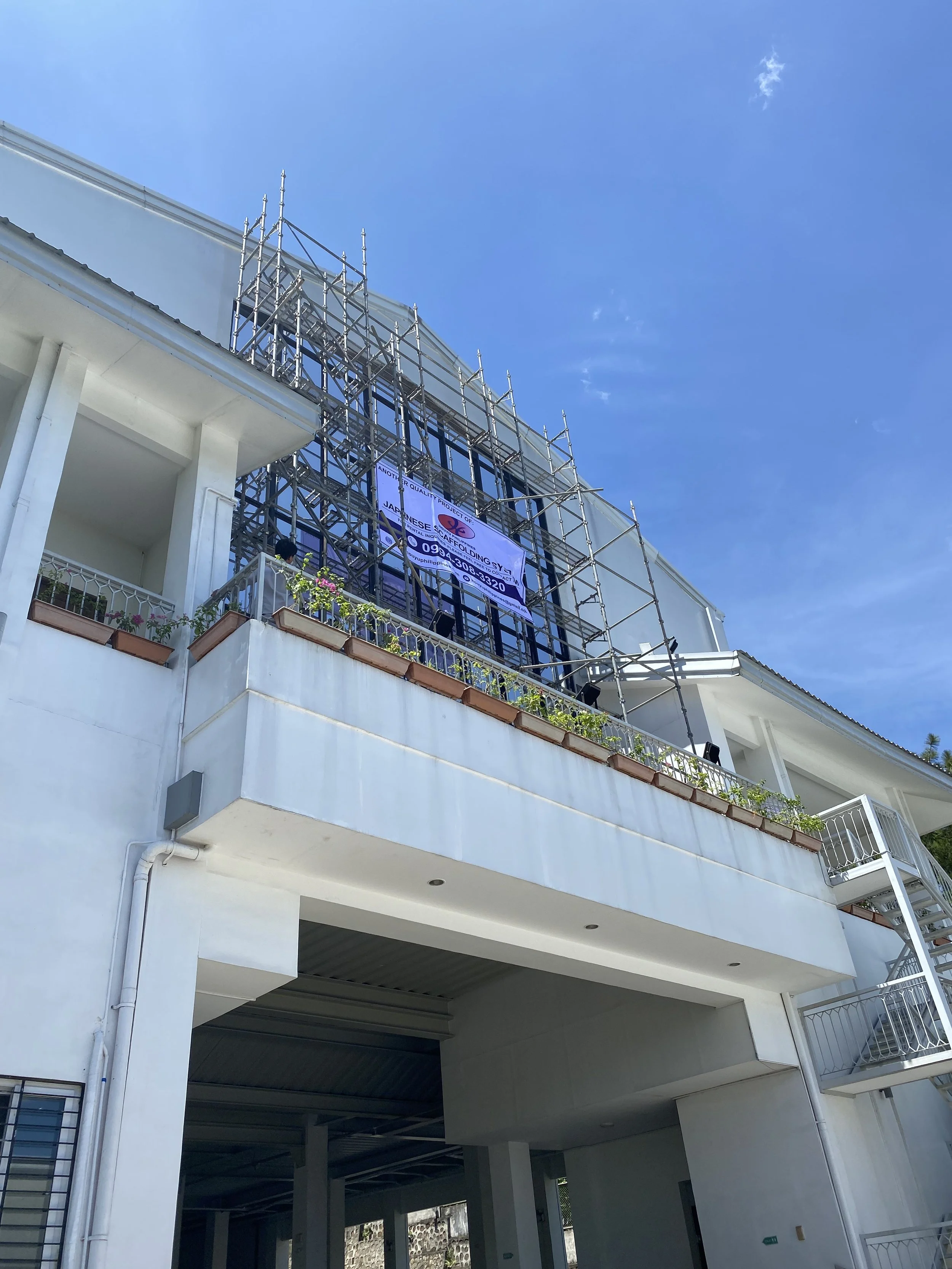 Construction scaffolding on a white multi-story building under a blue sky, with a balcony that has potted plants and a signage for a Japanese roofing company.