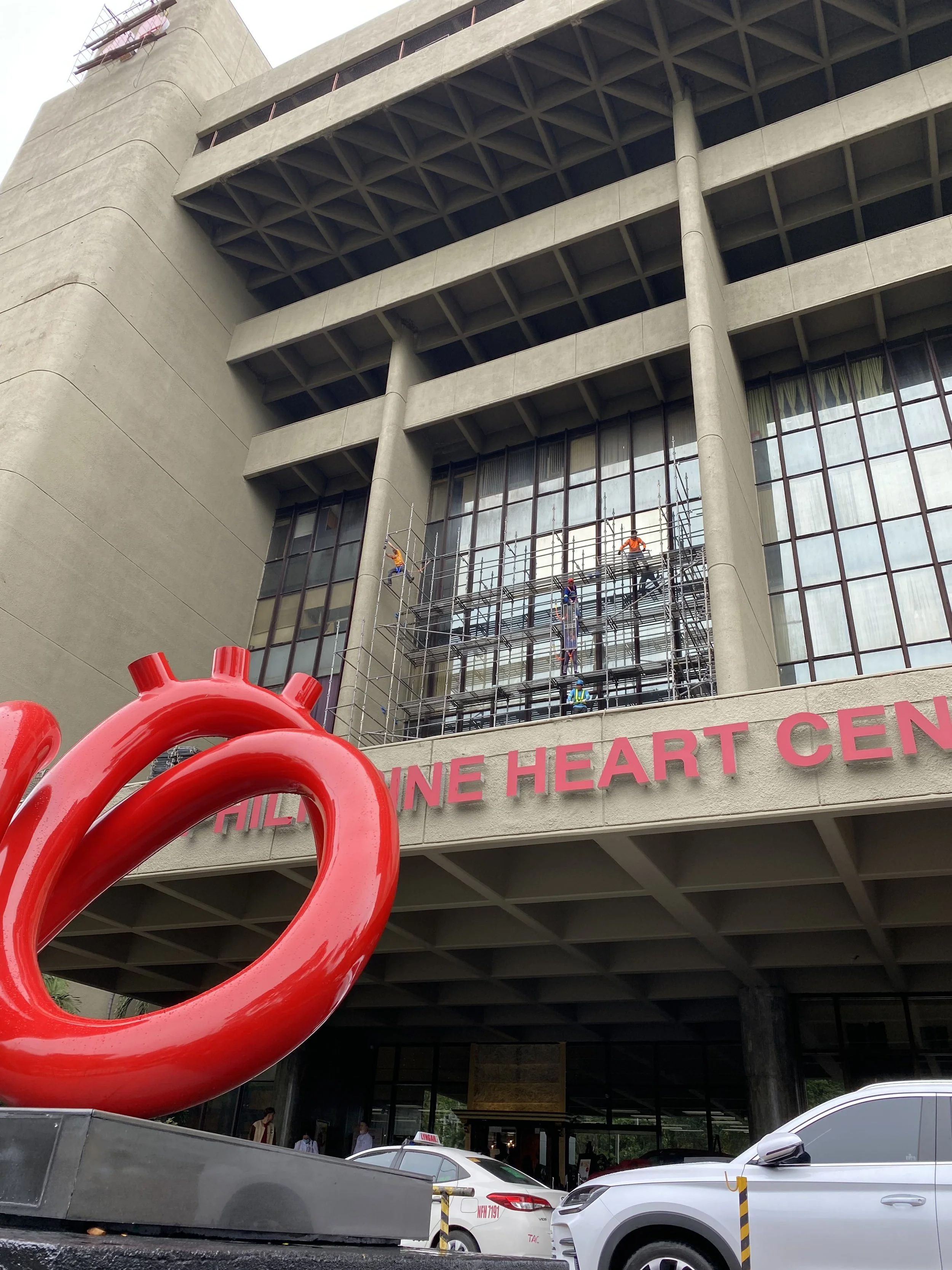 Workers on scaffolding repair the facade of a large modern building with glass windows. A large red heart sculpture sits in front of the building, which has a sign reading 'CHILINE HEART CENTER' or similar.