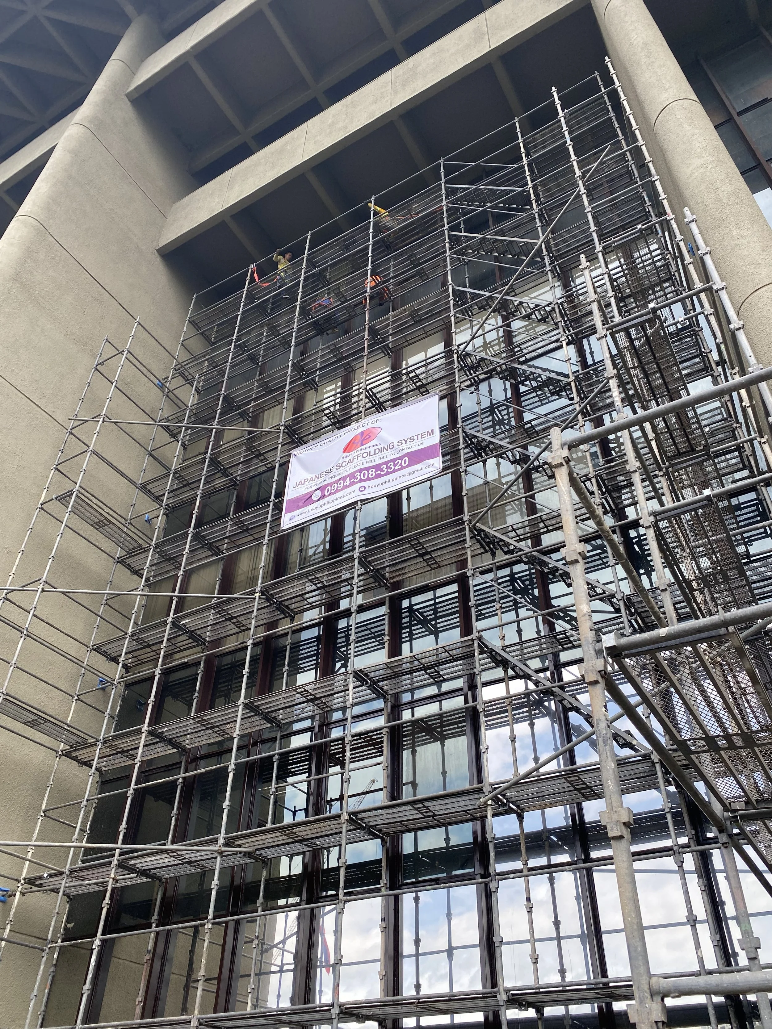 Scaffolding set up on a modern building with large glass windows, and a banner advertising Japanese scaffolding system with contact information.
