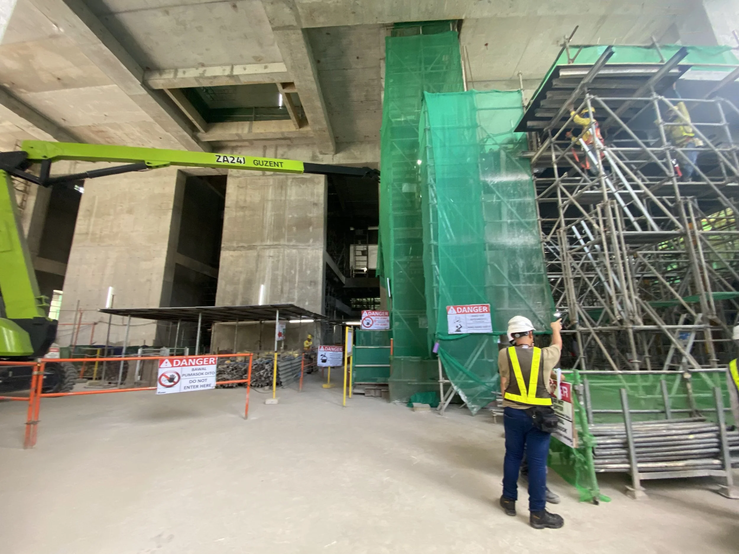 Construction site with scaffolding, safety signs, and workers in hard hats and vests, under an unfinished concrete structure.
