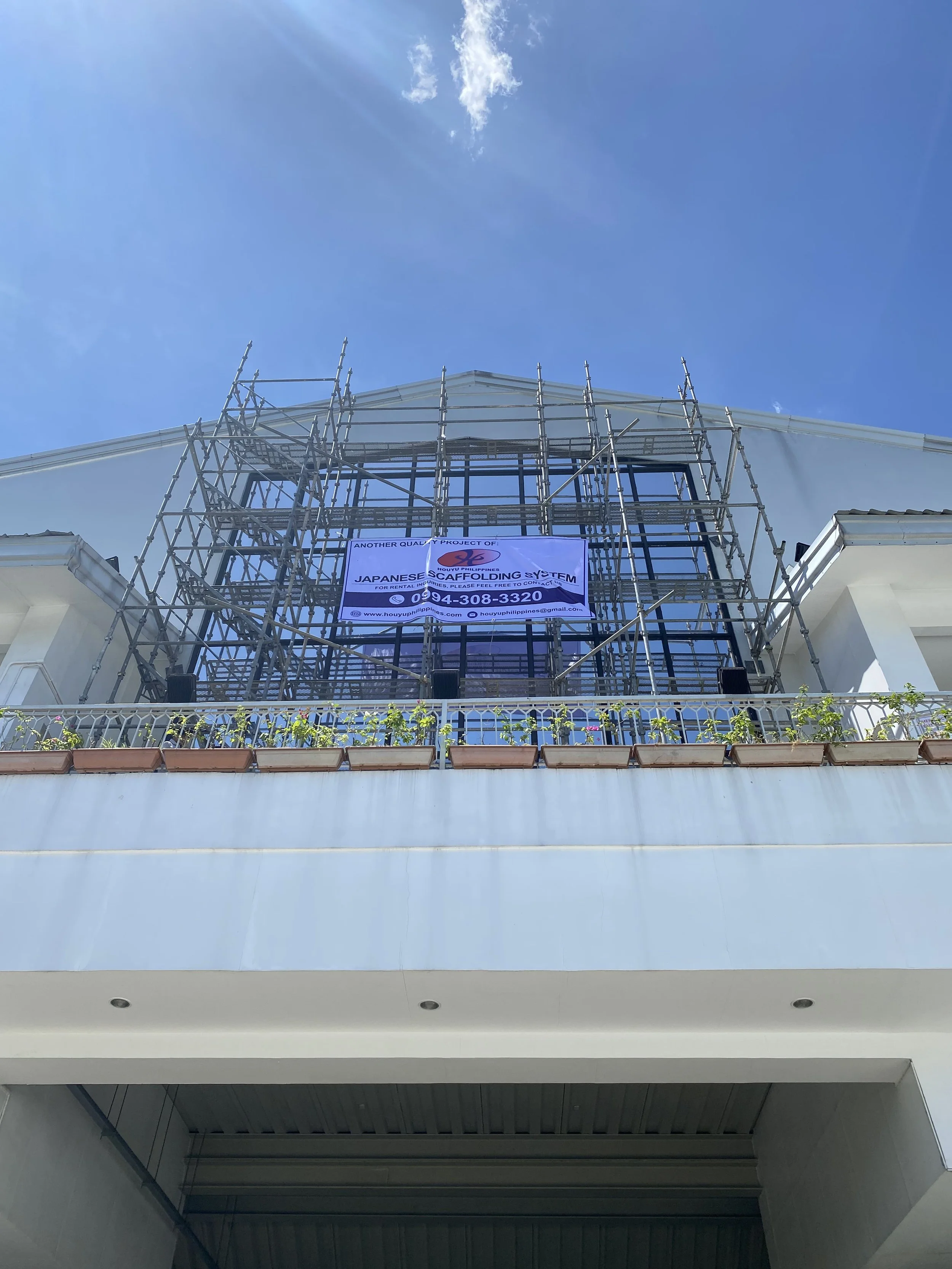A building under construction with scaffolding on the front and a sign that reads 'Japanese Folding System' and a contact number, against a clear blue sky.