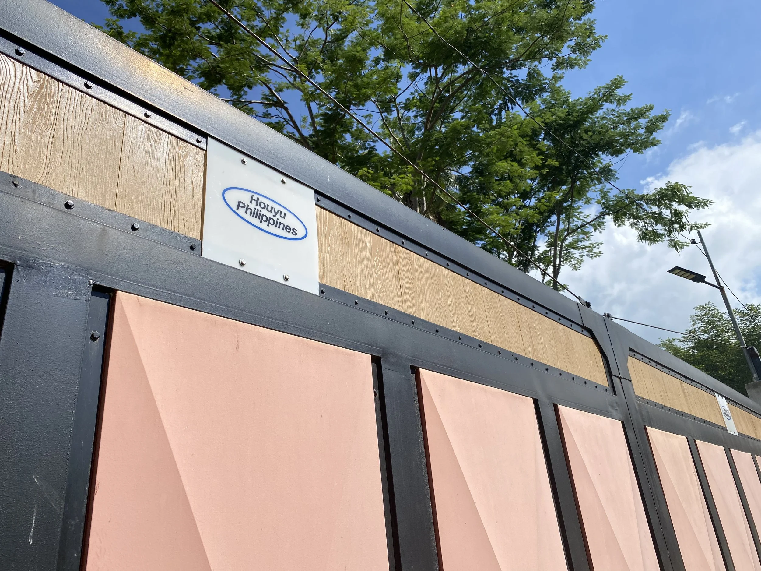 A fence with black metal frame, pink panels, and wooden upper section, with a small sign that says "Holuyu Philippines." In the background, there are trees, a streetlight, and blue sky with some clouds.