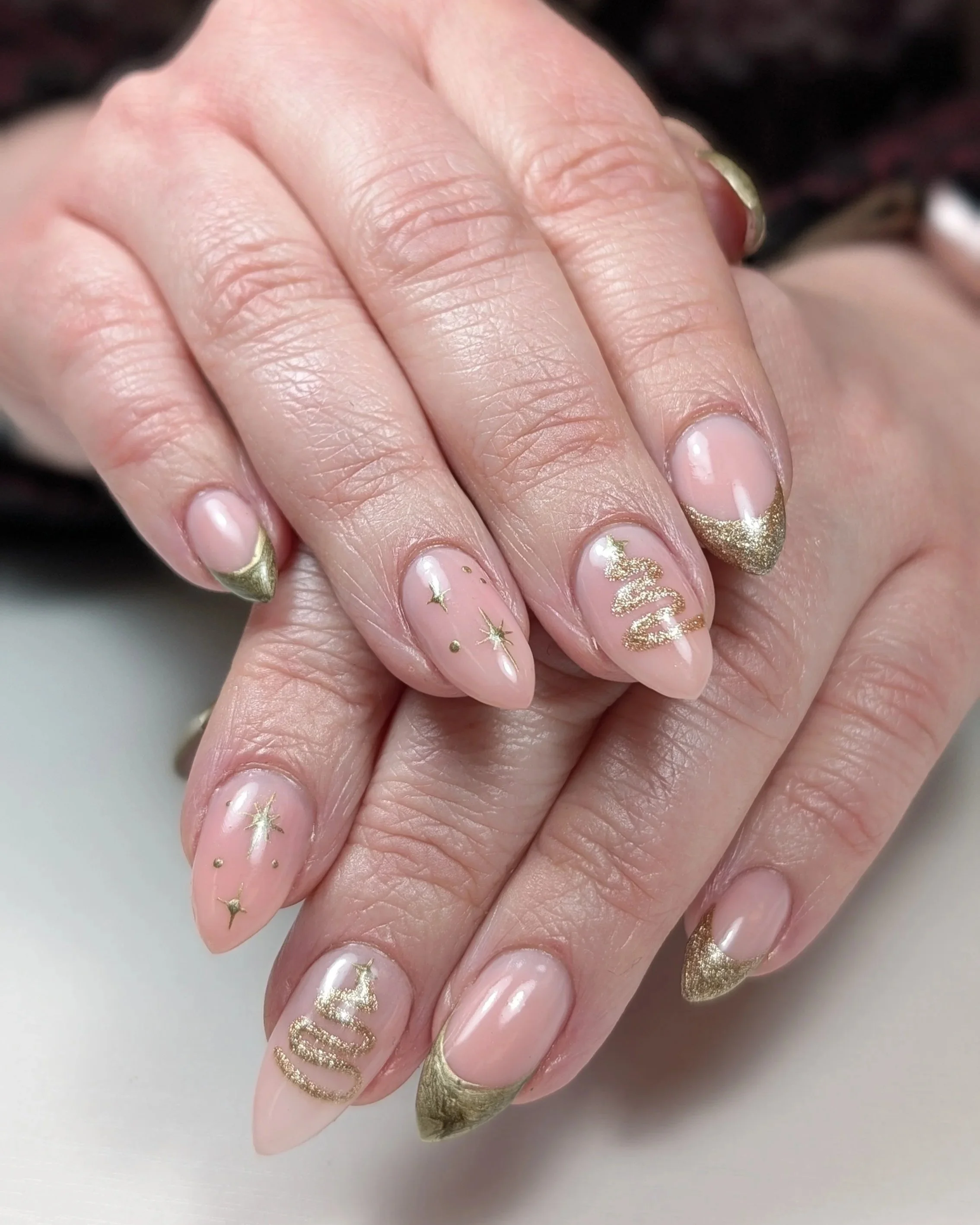 Hands with almond-shaped nude nails featuring gold French tips, small gold star accents, and metallic detailing.