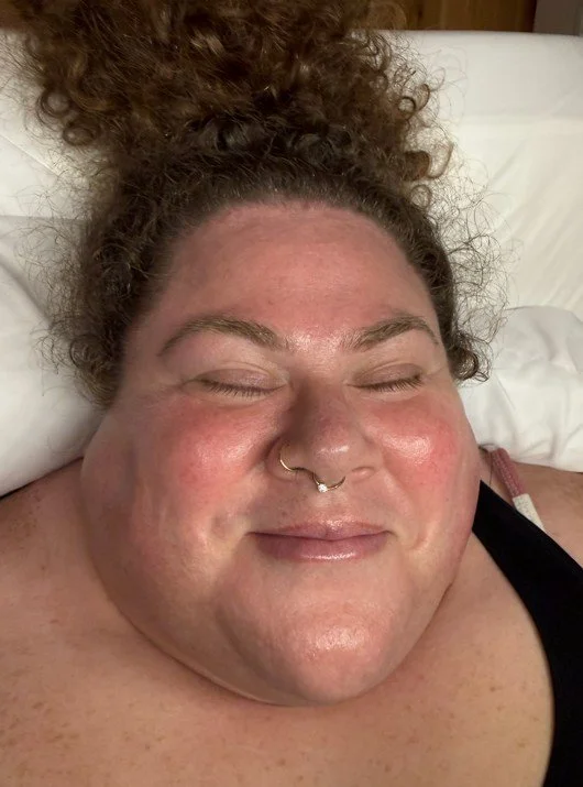 A client lying back with eyes closed, smiling softly, with flushed glowing skin and a nose ring, post-facial glow visible.