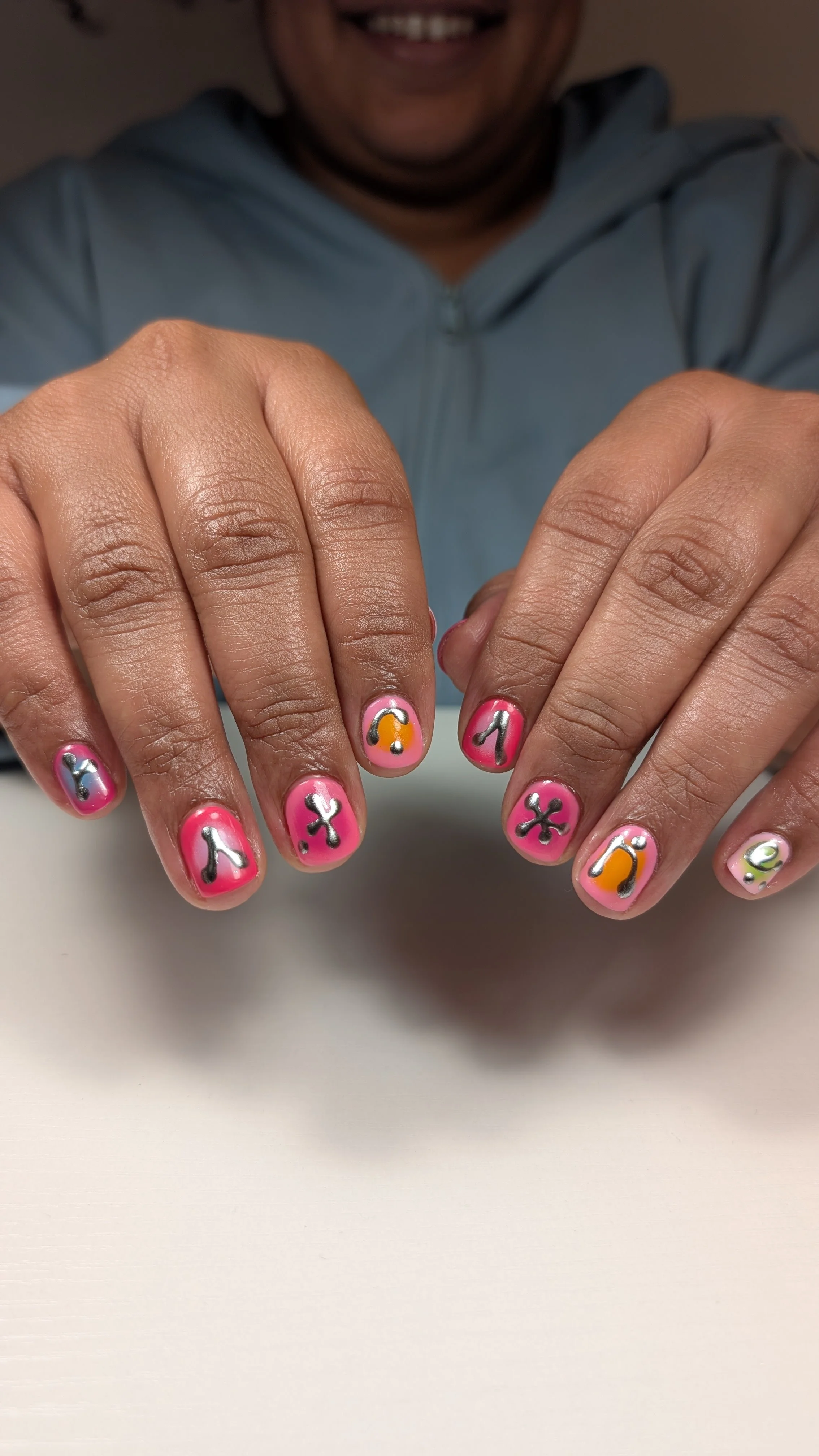 Hands with short pink nails featuring bold graphic designs in black, silver, orange, and green, including abstract symbols and raised metallic accents.