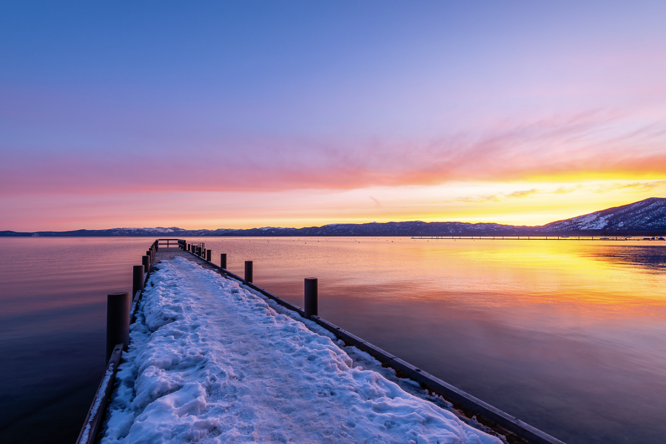 Snow-covered pier extending into a calm body of water at sunset, with colorful sky and mountain range in the background.