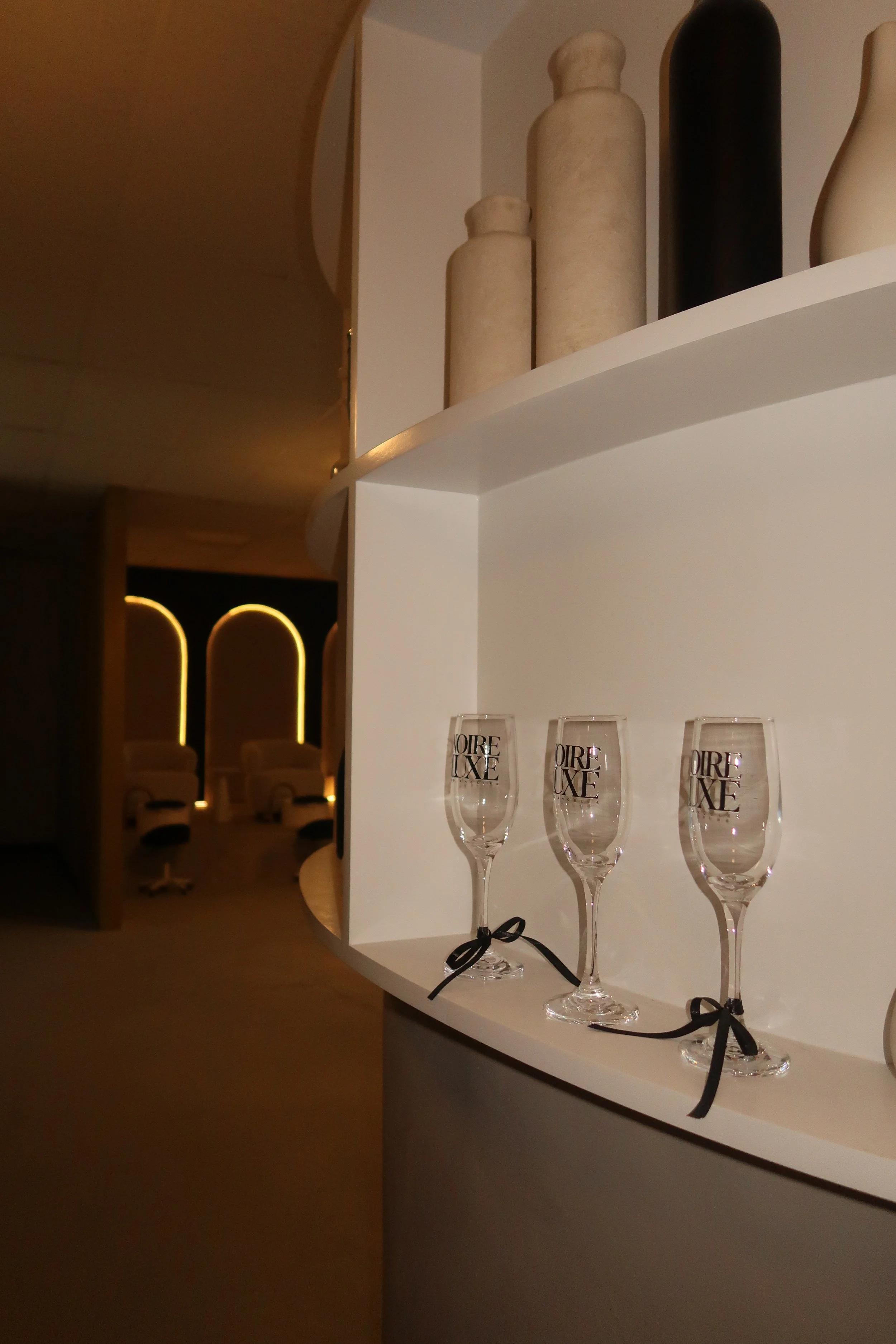 Three empty champagne glasses with black ribbons tied around the stems, placed on a white curved shelf. The glasses have the word QIRE LUXE printed on them.