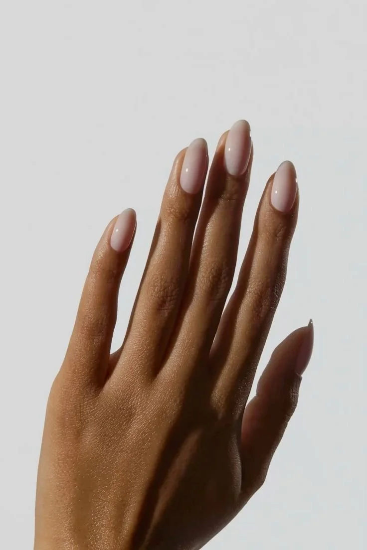 A person's hand with manicured nails painted in a nude or light pink color, against a plain light background.