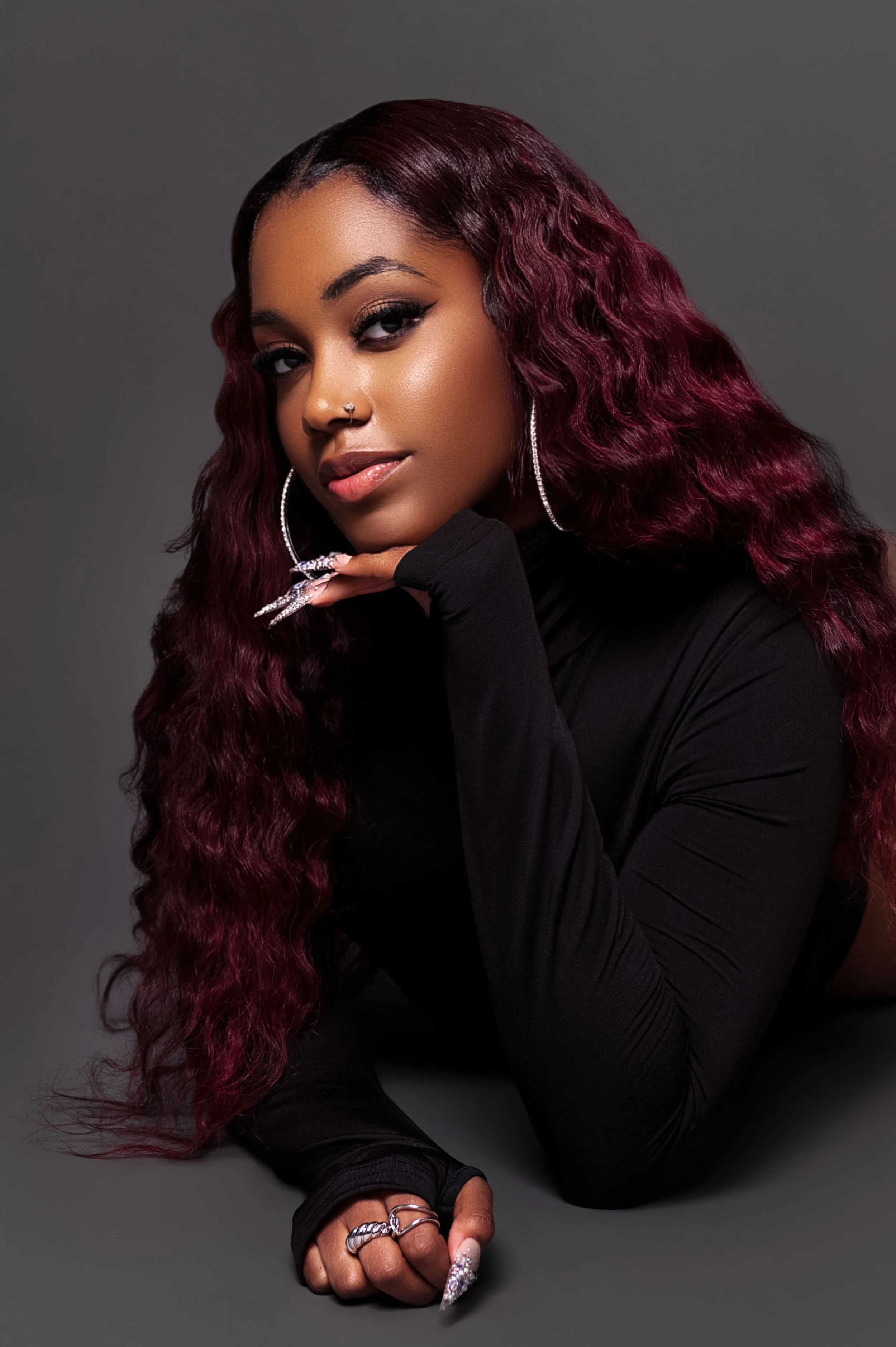 Portrait of a woman with long, wavy burgundy hair, wearing hoop earrings, a nose piercing, and rings, dressed in a black long-sleeve top, posing with her chin resting on her hand against a gray background.