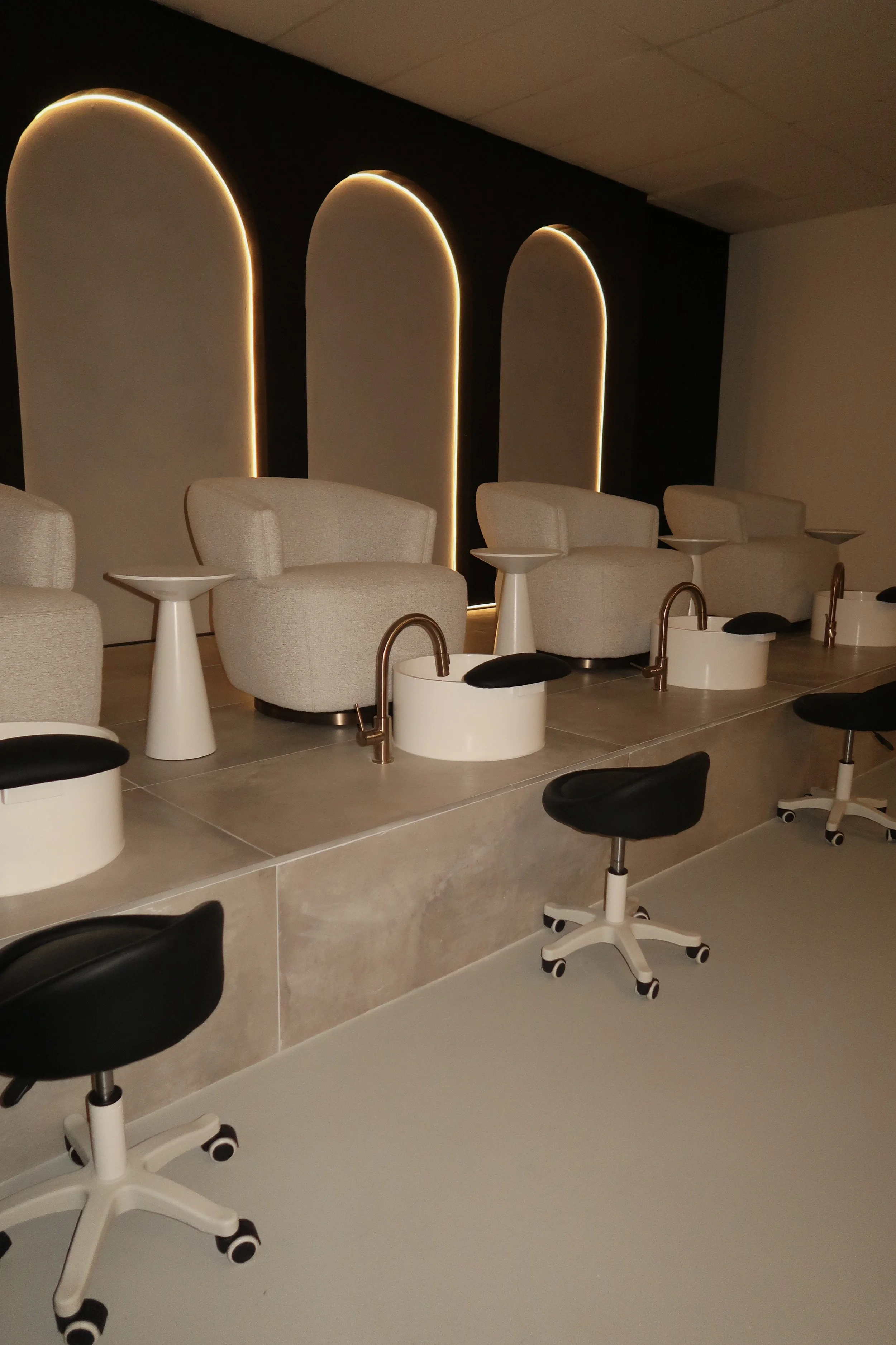 Nail salon chairs with foot baths and stools in a modern interior with backlit arches on a black wall