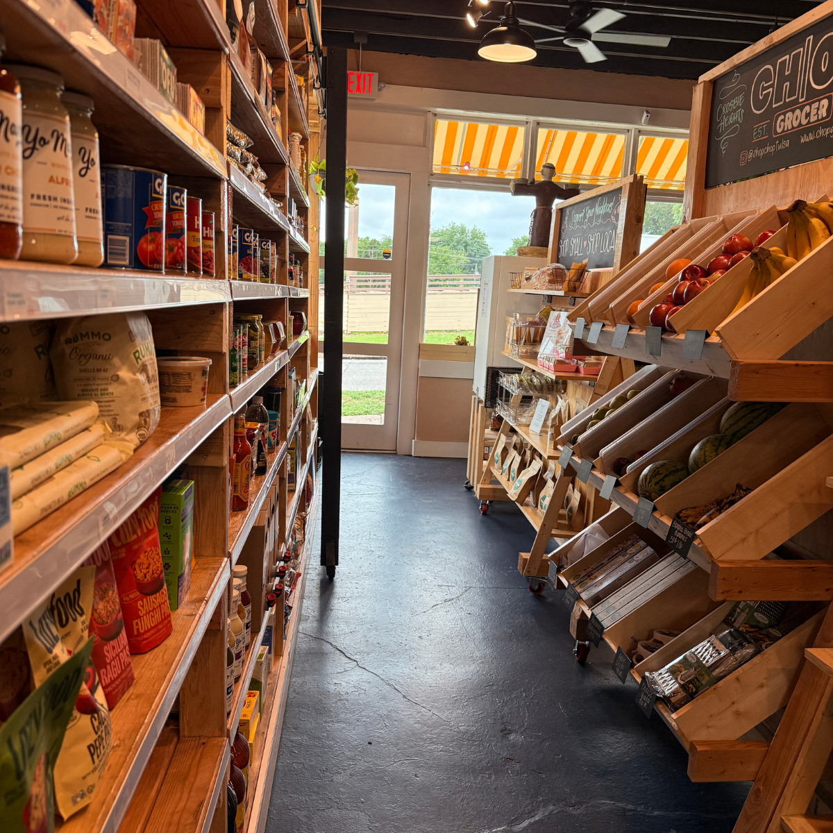 Inside a small grocery store with shelves stocked with canned goods, packaged foods, and fresh produce like apples, bananas, and watermelons. The store has a door and windows with yellow and white striped awnings outside, natural light, and a black ceiling with fans.