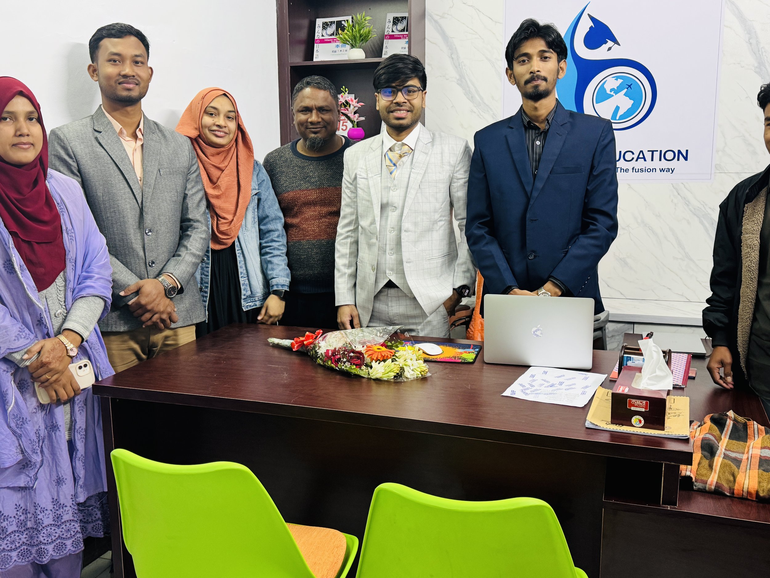 Group of seven people standing behind a table in an office or conference room, with a floral bouquet on the table and a laptop, papers, and office supplies. There is a white wall with a logo and a bookshelf with green plants and books in the backgrou