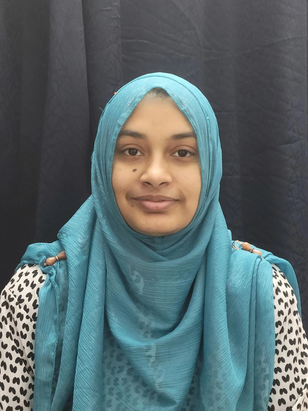 A woman with a light brown complexion wearing a blue hijab and a black and white dotted top, standing against a black background.