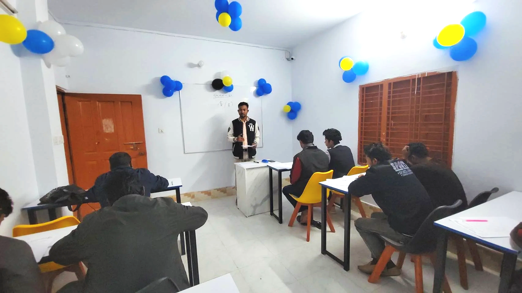 A classroom with students sitting at desks, facing a teacher standing at the whiteboard. The room is decorated with blue, yellow, and black balloons.