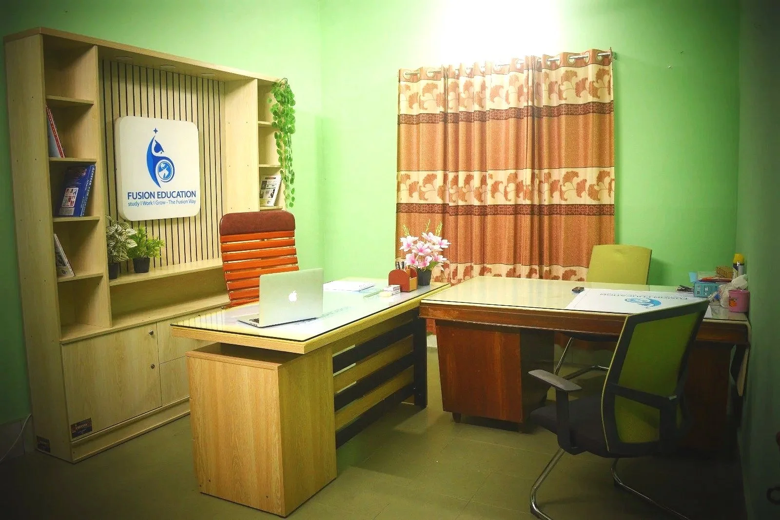 Office room with green walls, two desks, a red and a yellow chair, a laptop, a pink flower arrangement, books, and a window with floral curtains.