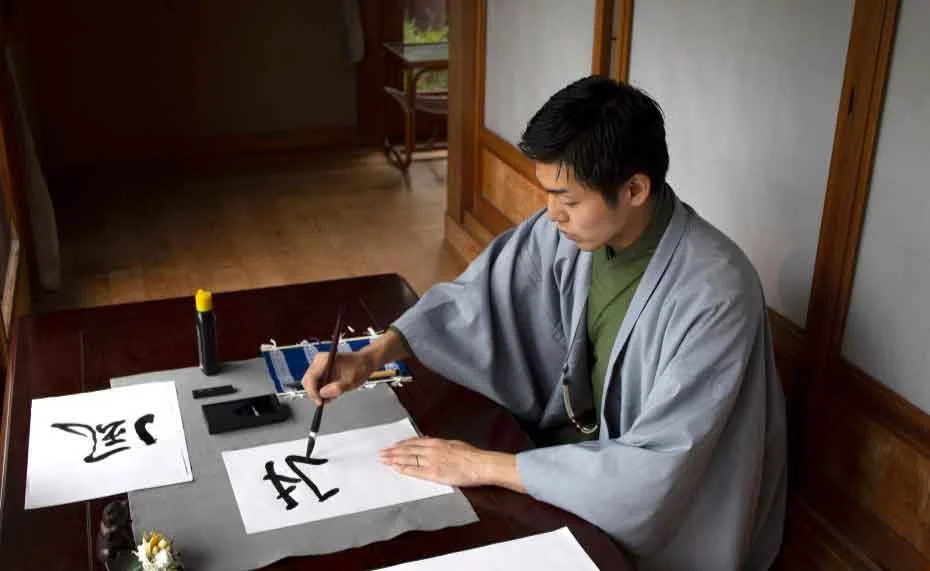 A person practicing traditional calligraphy at a wooden table with brush and ink, surrounded by paper sheets.