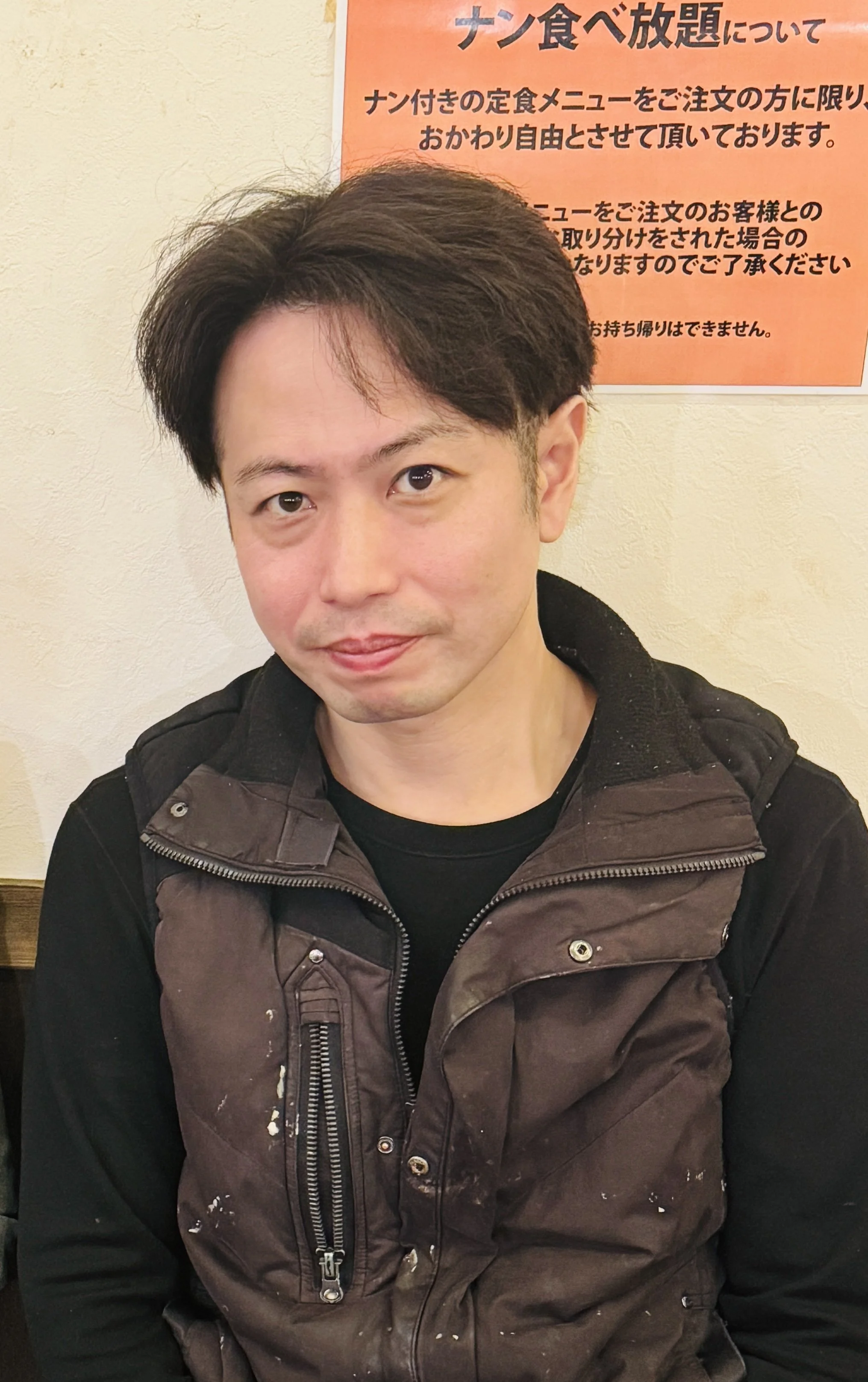 A man with dark brown hair wearing a black and brown jacket, sitting indoors near a wall with a Japanese sign.