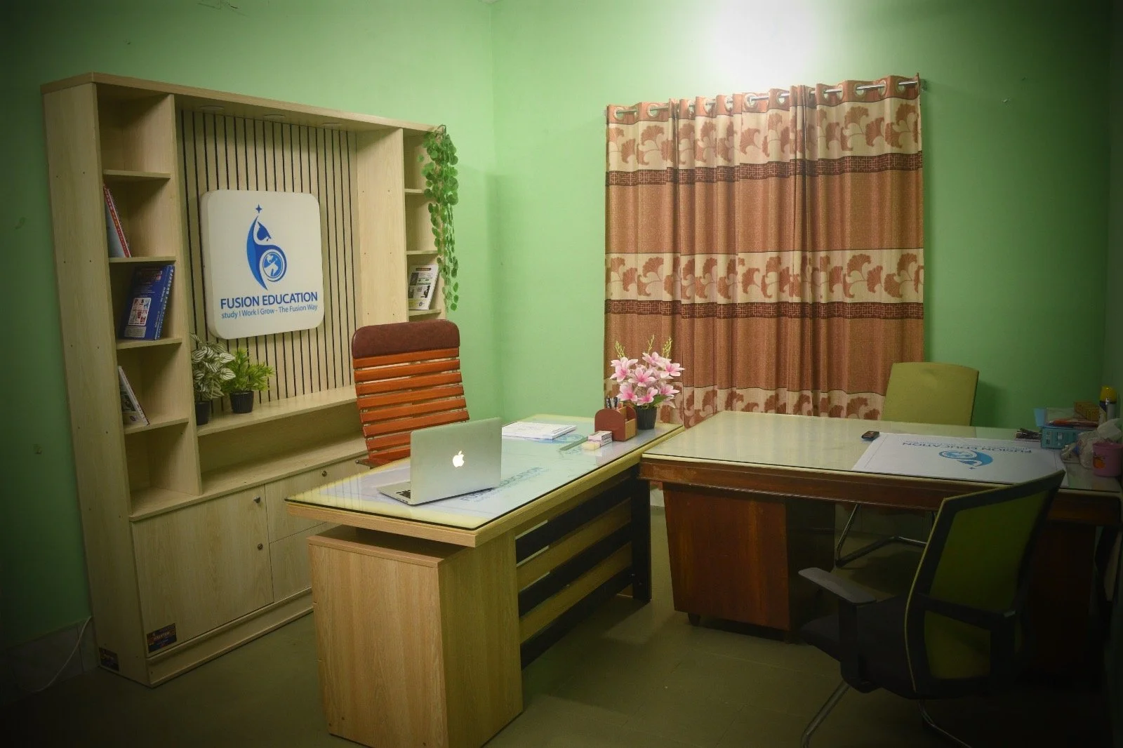 Office room with a green wall, wooden shelves, a desk with an Apple laptop, a bouquet of pink flowers, and curtains with a pink and beige pattern.