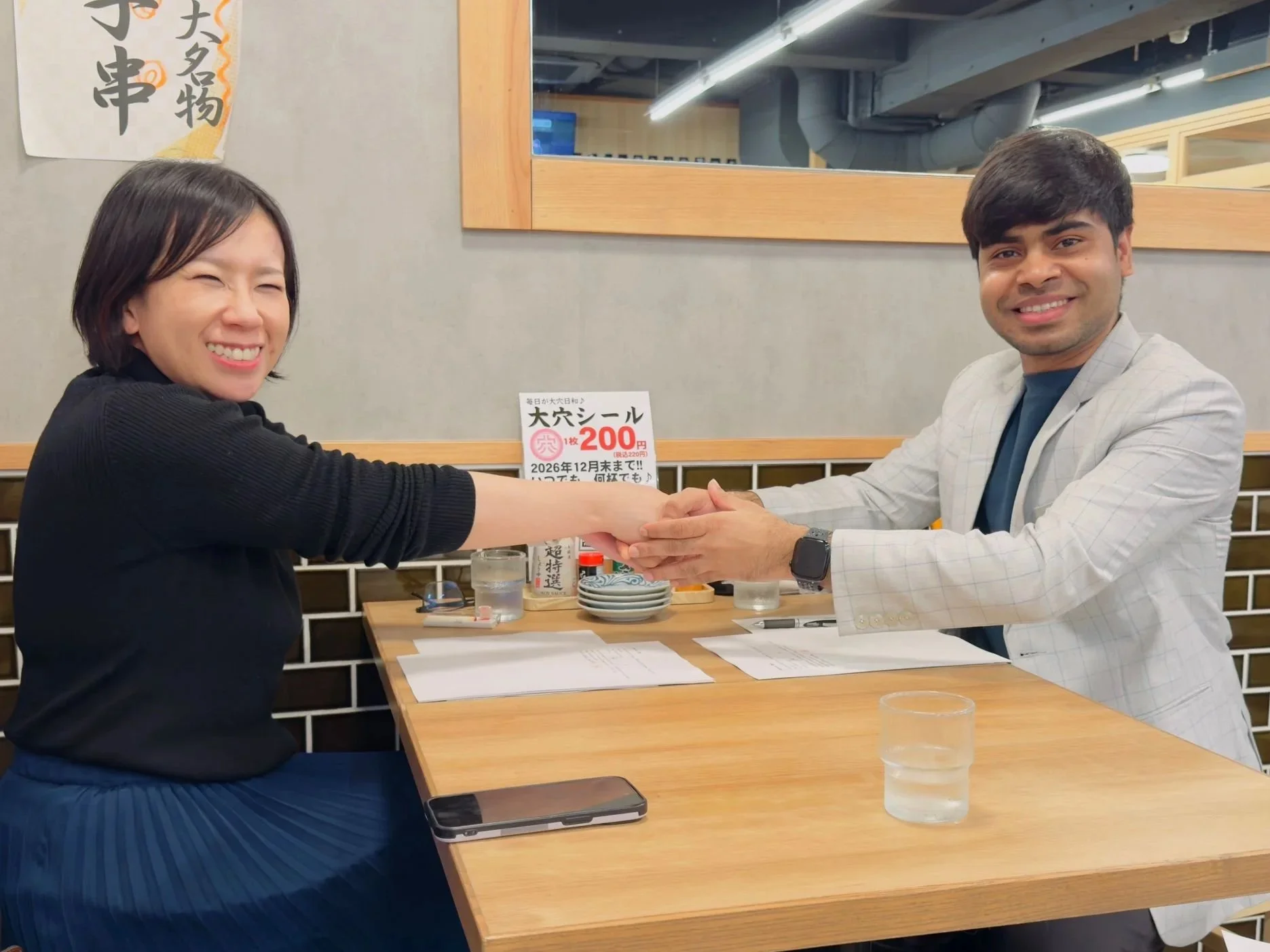 Two people, a woman and a man, smiling and holding hands across a wooden table in a restaurant. The woman has shoulder-length black hair and is wearing a black top and blue pants, while the man has dark hair and is dressed in a light-colored blazer over a blue shirt. There are papers, a glass of water, and a smartphone on the table, with a red and white sign, plates, and glasses visible in the background.