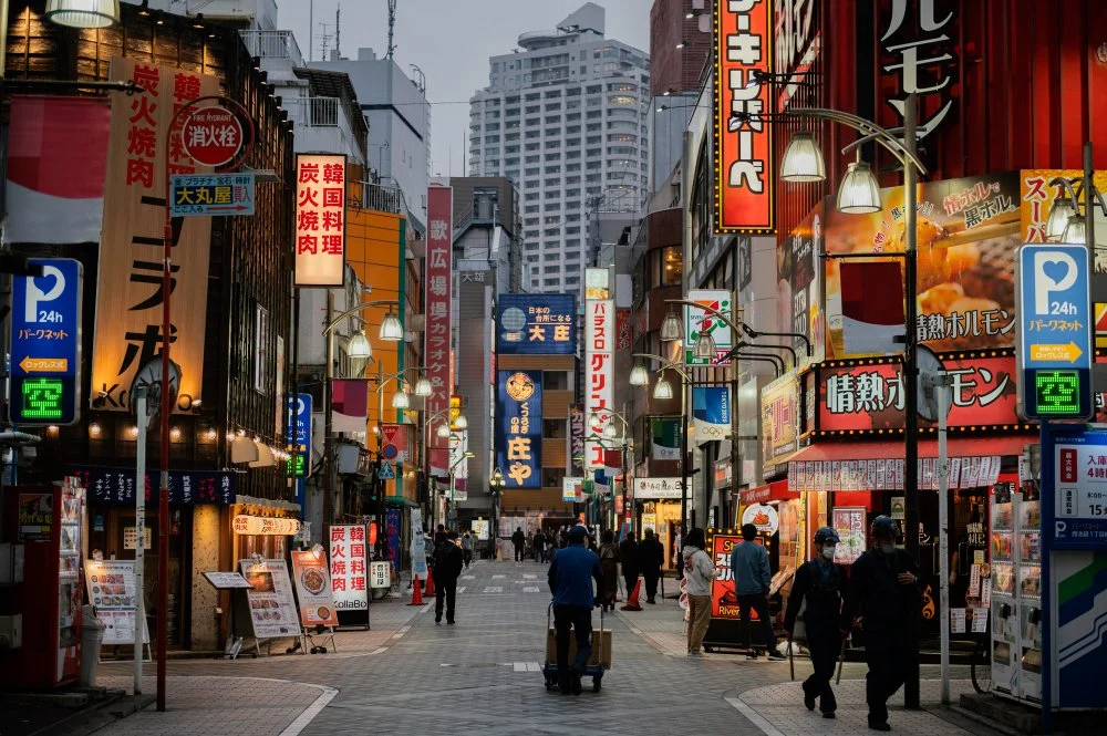 A busy city street in Japan with neon signs, storefronts, and pedestrians, some wearing masks, in the evening.