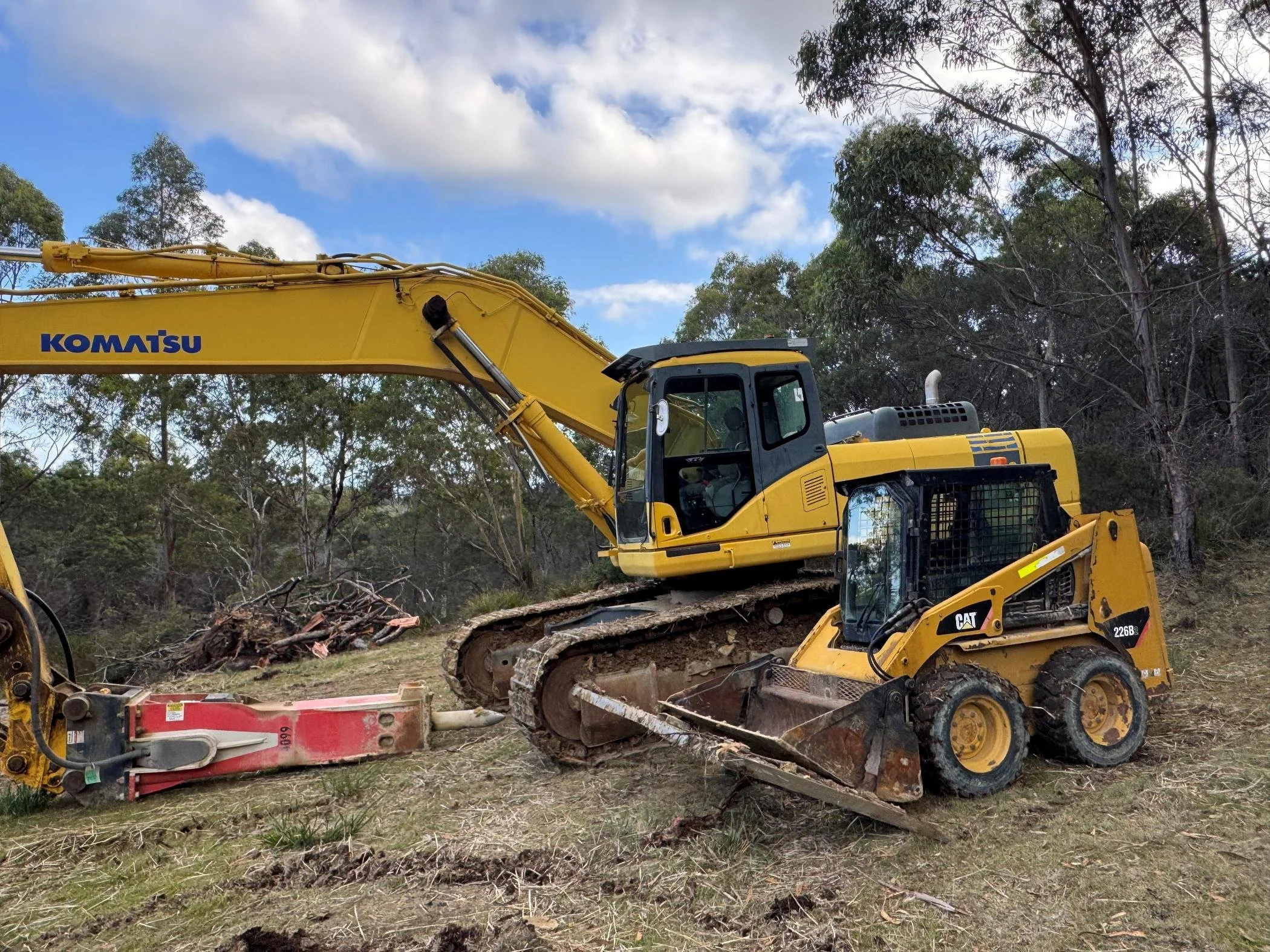 A yellow Komatsu excavator working on land clearing in a wooded area with trees and a partly cloudy sky.